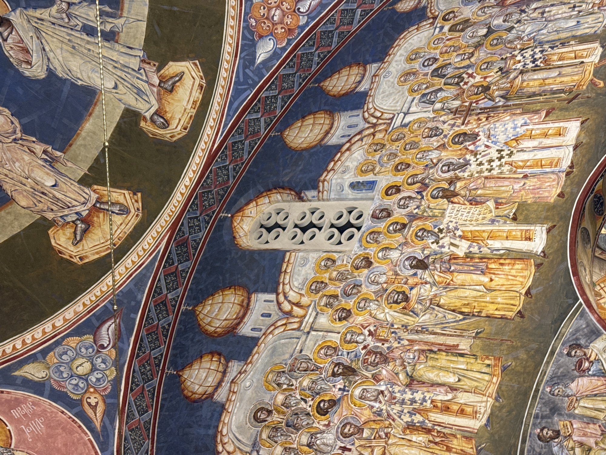 This image depicts a section of a fresco found in the Royal Church of St. Nicholas in Bar, Montenegro. The scene is part of a larger depiction of figures, presumably saints or biblical figures, rendered in traditional Byzantine style. The figures are characterized by halos, elongated bodies, and rich clothing. A large gathering of figures is shown to the right, while two singular figures appear on the left. Decorative motifs such as geometric patterns and floral designs frame the subjects and reinforce the architectural lines of the church. The fresco is executed in a muted color palette, dominated by blues, browns, and gold.