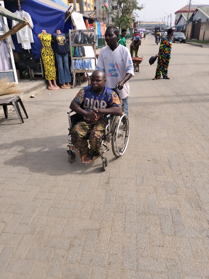 A daytime street scene in Alaiyabiagba, Nigeria, depicts a dark-skinned male in a blue "DIOR" t-shirt and camouflage trousers seated in a manual wheelchair. His bare feet rest on the footrests, and his hands are clasped in front. Another dark-skinned male, wearing a white t-shirt with a blue graphic, stands behind him, pushing the wheelchair along a light-colored paved sidewalk. To their immediate left, a shopfront displays clothing on mannequins, including a yellow animal-print maxi dress. The asphalt road to the right features pedestrians and vehicles in the mid-ground and distance, including a person in a colorful patterned jumpsuit holding a dark object and another pushing a handcart. Commercial buildings line the street.