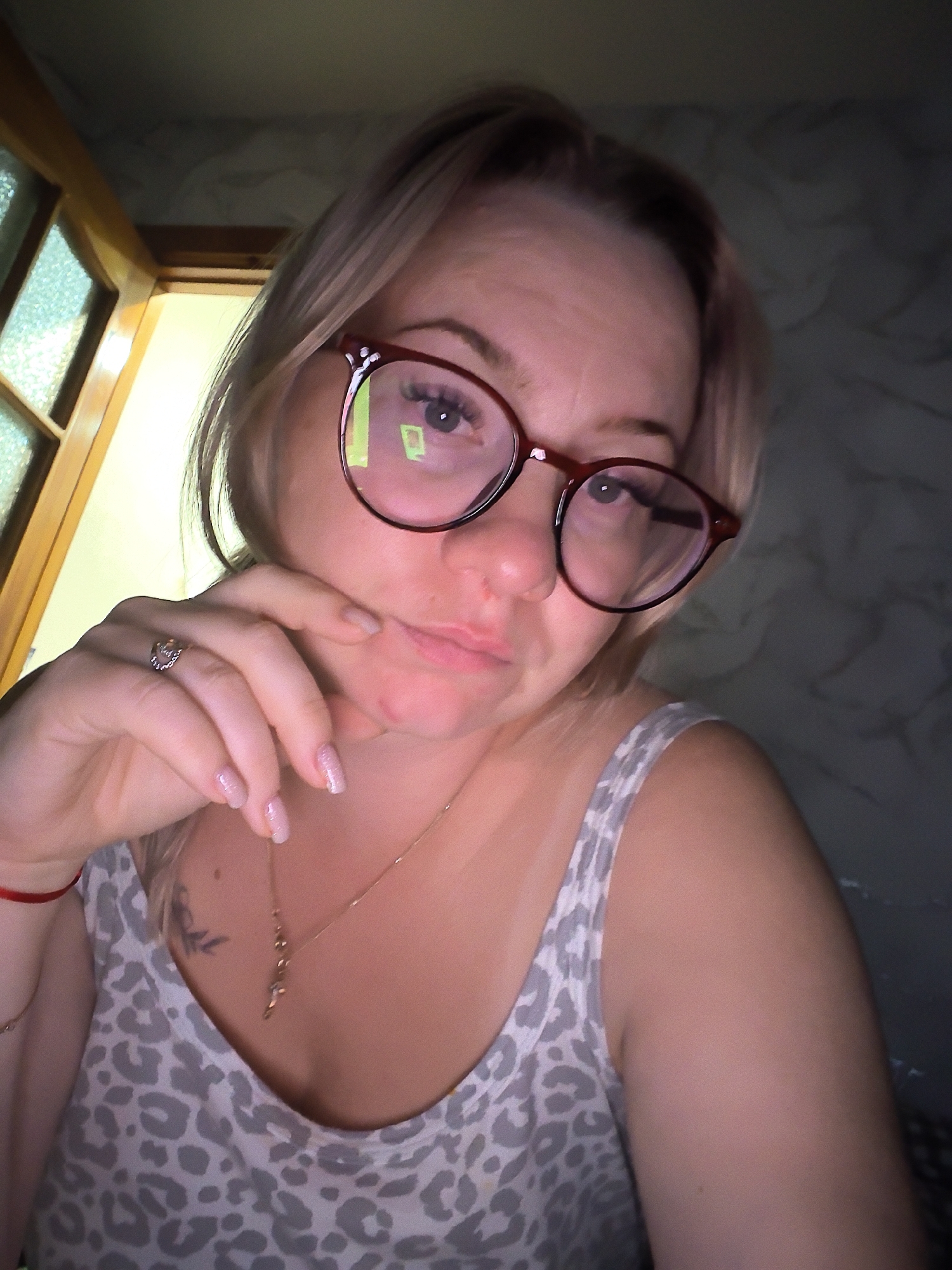 A woman with blonde hair and red-rimmed glasses is taking a selfie. She is wearing a grey and white leopard-print tank top and a gold cross necklace. Her right hand is resting on her chin, and she is wearing a ring on her ring finger. A red string bracelet is visible on her left wrist. A tattoo is visible on her chest. The background shows a wooden door frame with glass panes and a textured wall. The lighting suggests an indoor setting, possibly a room in Chaikovsky, Russia.