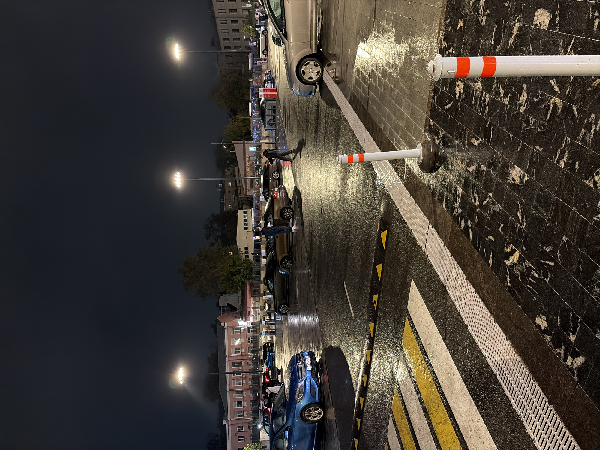 The image depicts a nighttime scene in a city, likely Moscow, Russia, judging by the architectural style of the buildings and the Cyrillic script visible on some signs. The foreground features a wet crosswalk with yellow and white stripes, and a drainage grate. Several cars are parked or driving on the wet asphalt surface. In the middle ground, a person is walking across the parking lot. The background shows a building with red accents and multiple windows, illuminated by streetlights. Two tall lampposts with multiple lights are visible, casting a bright glow on the scene. The overall environment is dark, wet, and urban.