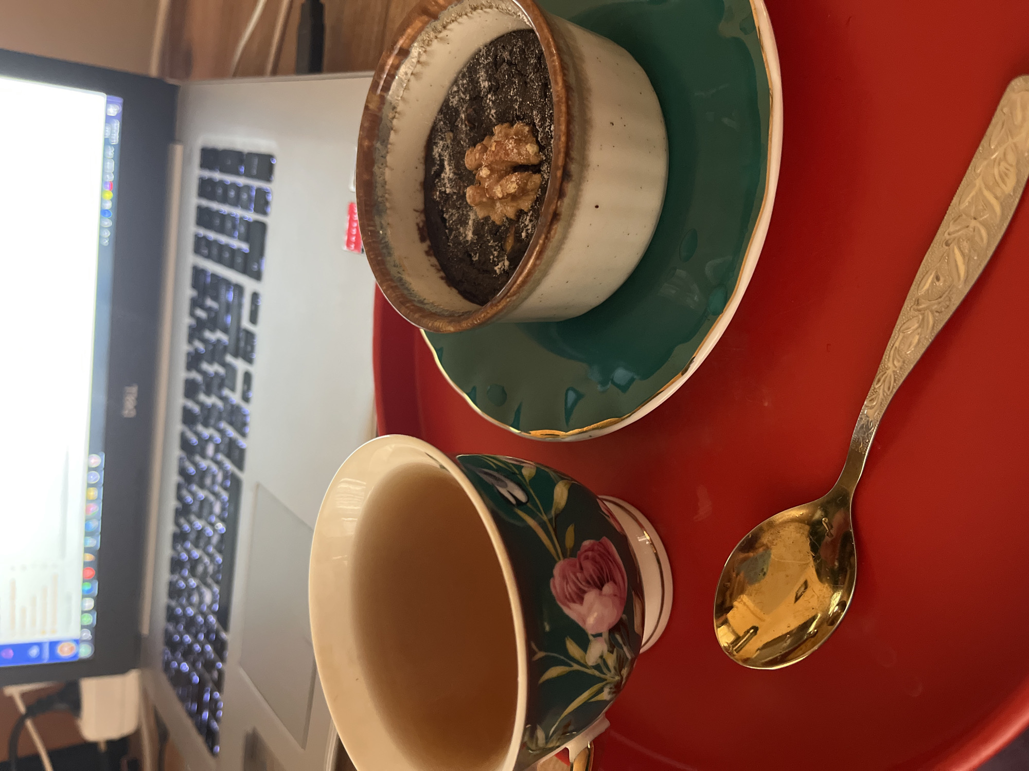 A cup of tea and a dessert are displayed on a red tray next to a laptop. The cup, decorated with floral patterns, contains a clear, light-colored beverage. The dessert, topped with a walnut, is presented in a small ceramic dish placed on a green saucer. A silver spoon lies on the tray beside the cup. The laptop, with its screen and keyboard visible, rests adjacent to the tray. The image does not show any people or any details that would indicate that this picture was taken in Alanya, Turkey.