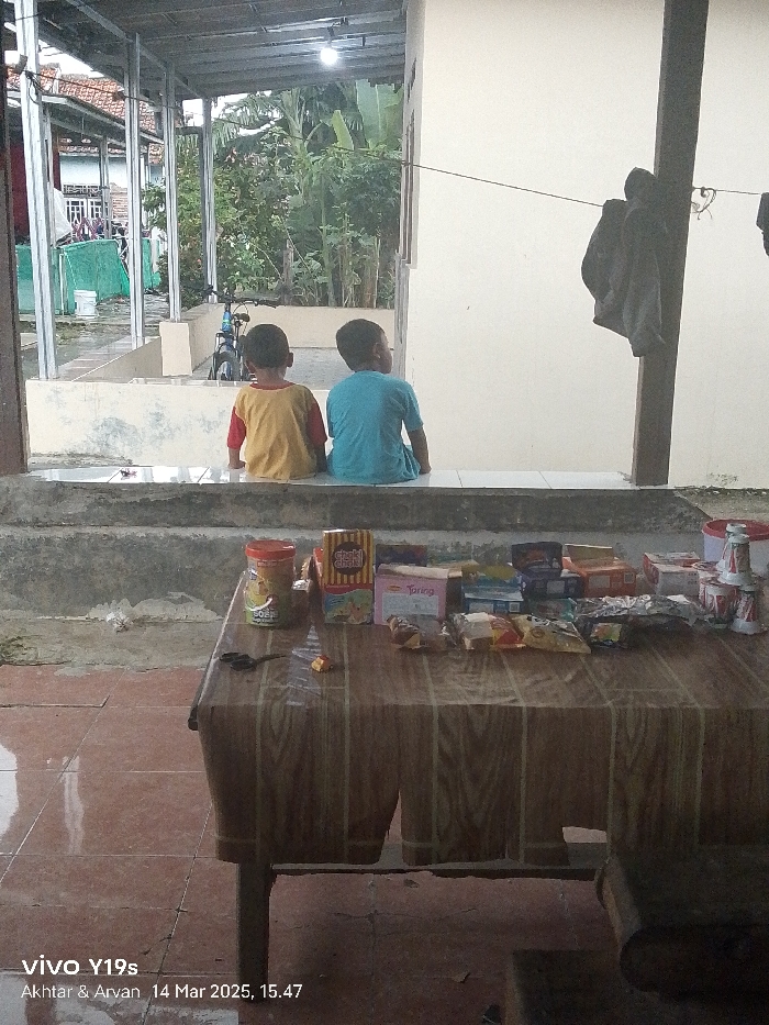 Two young boys are sitting on a concrete ledge outside a home in Anjatan, Indonesia (latitude: -6.419, longitude: 107.978). The boys are facing away from the camera, and the one on the left is wearing a yellow shirt, while the one on the right is wearing a blue shirt. They are looking at something off-camera, and there is a table in front of them. The table is covered with a brown tablecloth and is laden with various packaged snacks and drinks. The scene is set in a partially covered outdoor area with a view of a backyard and a partly obscured bicycle. The floor is a mixture of concrete and tiles. The atmosphere is relaxed and casual. A single brown jacket is hanging on a post in the background. The image was taken on March 14, 2023, at 15:47.