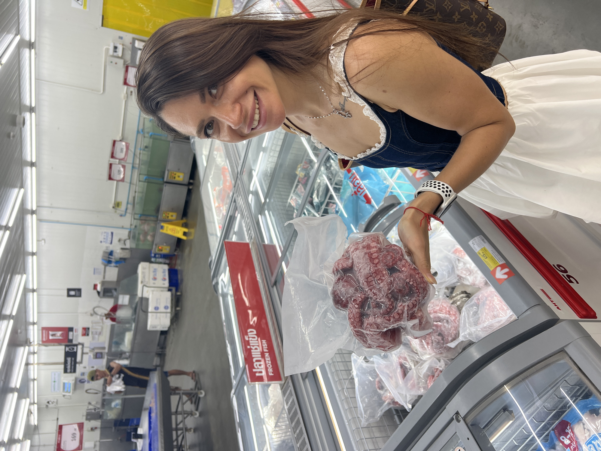 The image depicts a woman in a supermarket aisle, potentially in Baan Hua Thanon, Thailand. She is holding a plastic bag containing frozen octopus. The aisle features refrigerated displays of frozen seafood, marked with signs in both Thai and English ("Frozen Fish"). The woman is smiling and wearing a denim-style top with lace detailing, a white skirt, and a watch. A Louis Vuitton handbag is partially visible over her shoulder. In the background, other shoppers and store staff can be seen. The overall environment suggests a modern supermarket setting.