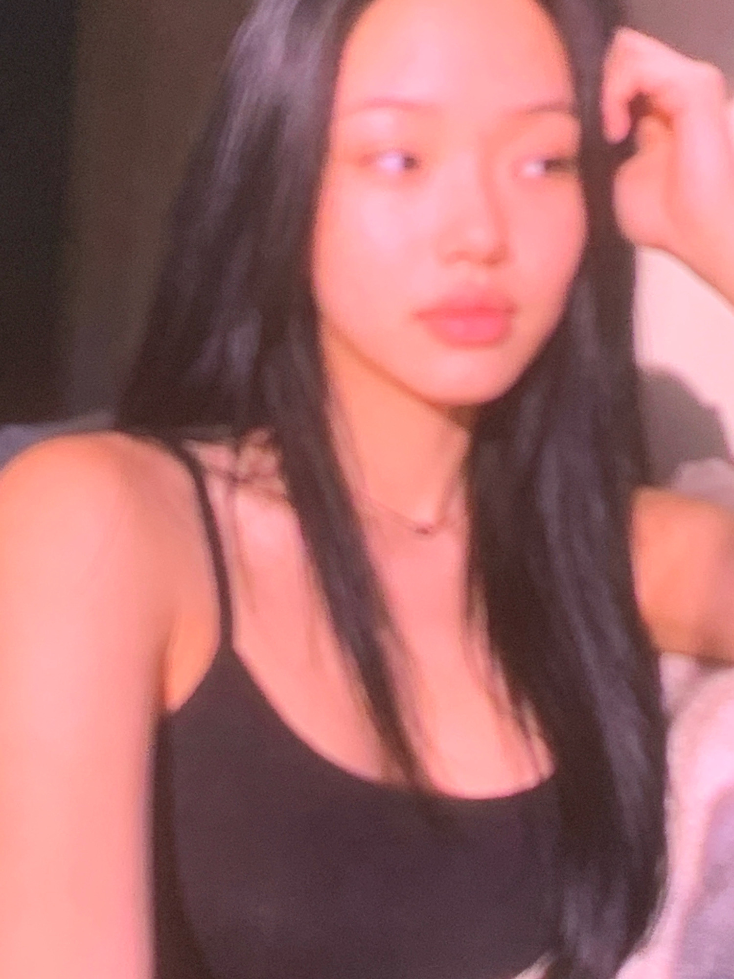 A close-up, portrait-style, slightly blurry photograph shows a young East Asian female from the chest up, against a dark background with warm lighting. She has long, dark hair that falls over her shoulders and appears to be wearing a black tank top. Her skin is fair and has a pinkish hue. Her head is turned to her right, and her eyes are looking in that direction. Her right hand is raised near her face, with her fingers touching her hair or head. A thin, dark necklace with a small pendant is visible on her décolletage. The overall mood is soft and intimate due to the lighting and the focus on her face. The location is not specified in the image.