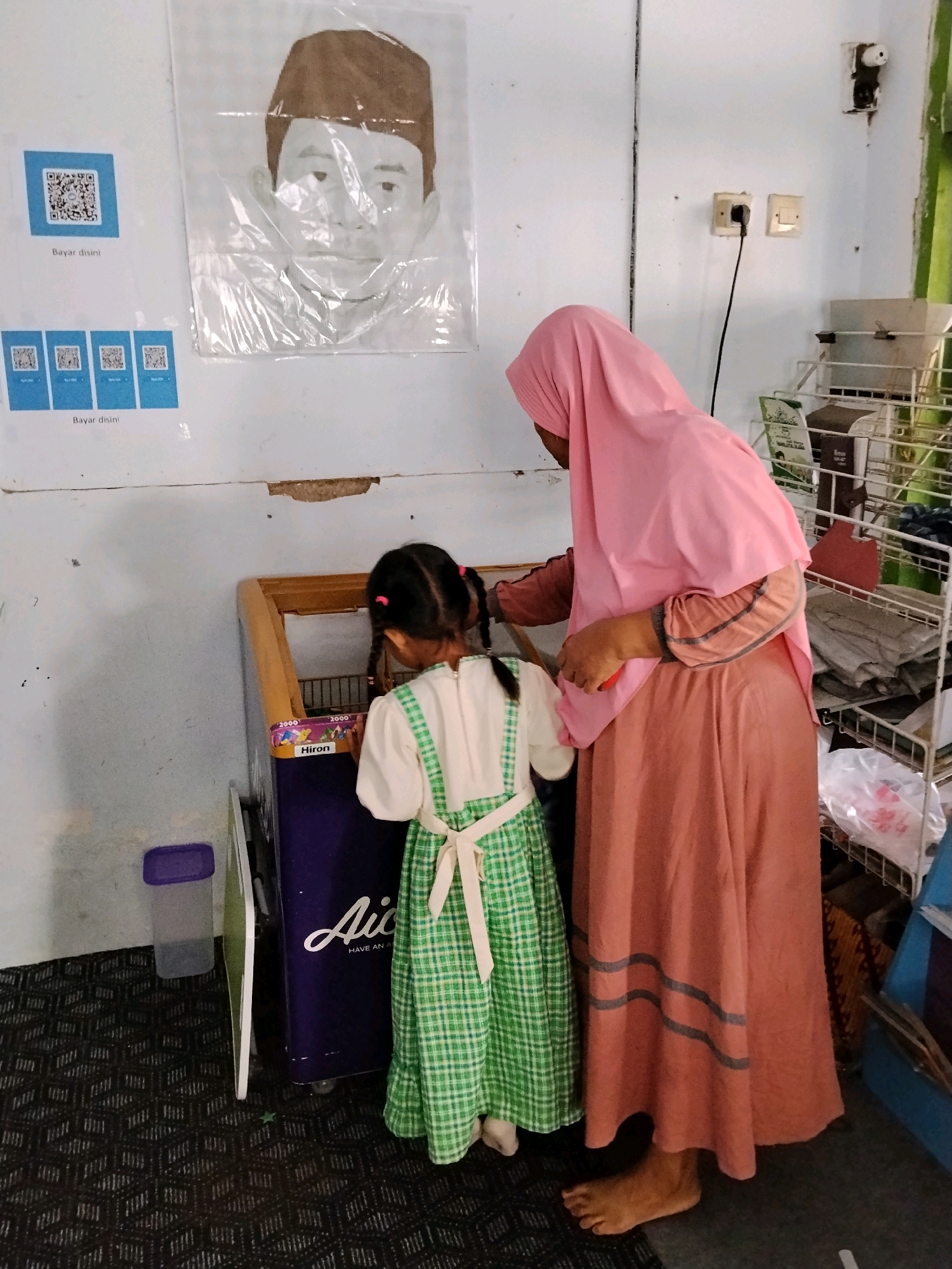 In the city of Panggungan, Indonesia, a young girl wearing a green and white checkered dress with pigtails is looking into a purple freezer chest. Beside her, a woman wearing a pink hijab and a long peach-colored dress is standing and observing. The freezer chest has text that reads "Hiron" and displays a colorful image of stars. On the wall behind them, a portrait of a person wearing a dark cap is displayed, along with several QR codes labeled "Bayar disini". Electrical outlets and a light switch are visible on the wall. A metal shelving unit with various items is positioned to the right. The floor is covered with a patterned carpet.