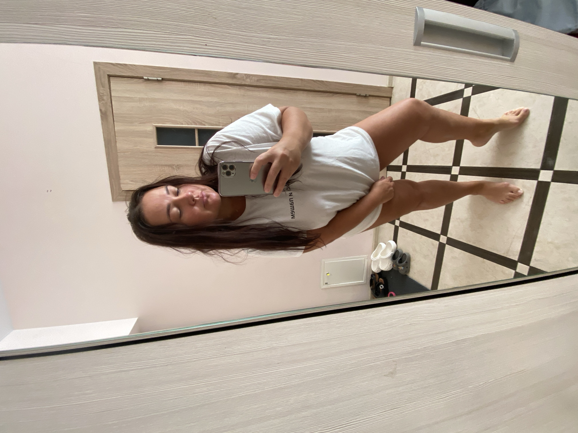 A woman is taking a selfie in a mirror, wearing a white t-shirt and no pants. She is standing in what appears to be a bedroom or dressing area with a tiled floor and a wooden door in the background. There are several pairs of shoes on the floor near the mirror, and a cabinet is visible to the left. The scene is indoors, and the lighting suggests it is daytime. There is no visible indication of the city of Sochi, Russia, or any other specific urban location.