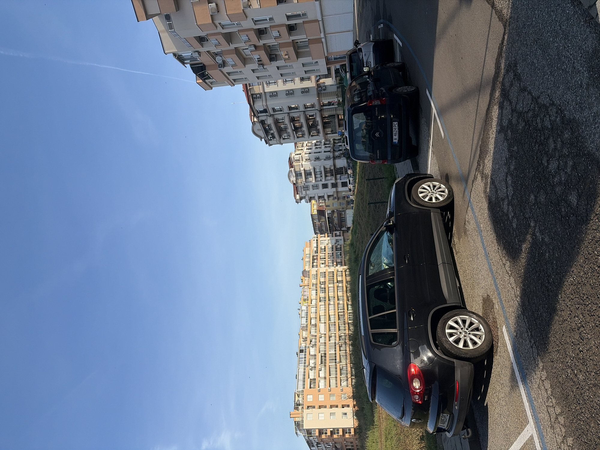 The image depicts a parking lot with several parked cars and apartment buildings in the background. The sky is blue and clear. A black Volkswagen Tiguan is parked along the right side of the image, and a dark-colored Citroen Nemo is parked further up the lot. Several other cars are parked behind the Citroen. The location appears to be an urban residential area in Unknown, Unknown. The apartment buildings are multi-story with balconies and a beige or light-yellow facade. The parking lot is marked with white lines.