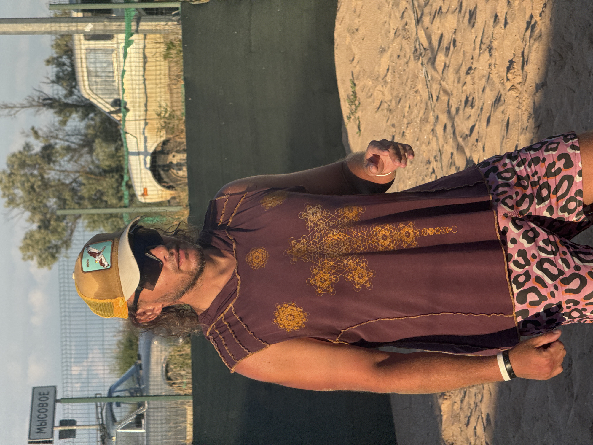A medium shot captures a man standing on a sandy beach. He is wearing a sleeveless hooded t-shirt with a geometric pattern and leopard-print shorts. A baseball cap with a patch is on his head, and he has sunglasses on. The man is looking to his right, with his left arm bent at the elbow and his hand outstretched. In the background, a fence separates the beach from a street with a sign that reads "МЫСОВОЕ," and two parked cars. Trees and a white vehicle are visible behind the fence. The sun is casting long shadows on the sand.