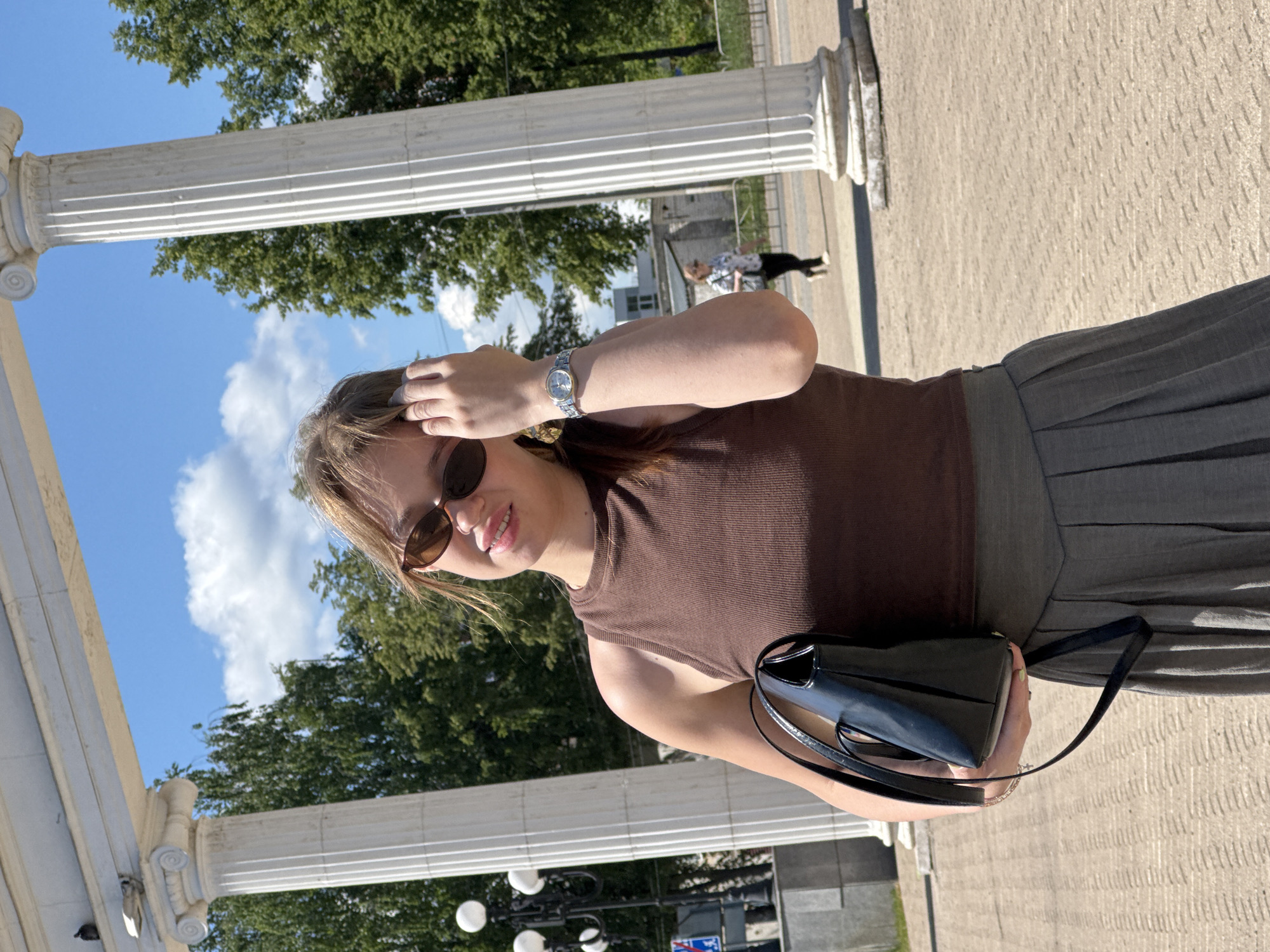 A young woman with light brown hair and sunglasses is posing in front of a white architectural structure with large columns. She is wearing a brown sleeveless top and a grey pleated skirt. She is holding a black purse in her left hand and her right hand is touching her hair. A silver watch is visible on her left wrist. In the background, there are trees, a blue sky with white clouds, and another person walking. The ground is paved with light-colored rectangular stones. The scene appears to be outdoors in a park or public square, likely in the city of Chaikovsky, Russia, given the architectural style.