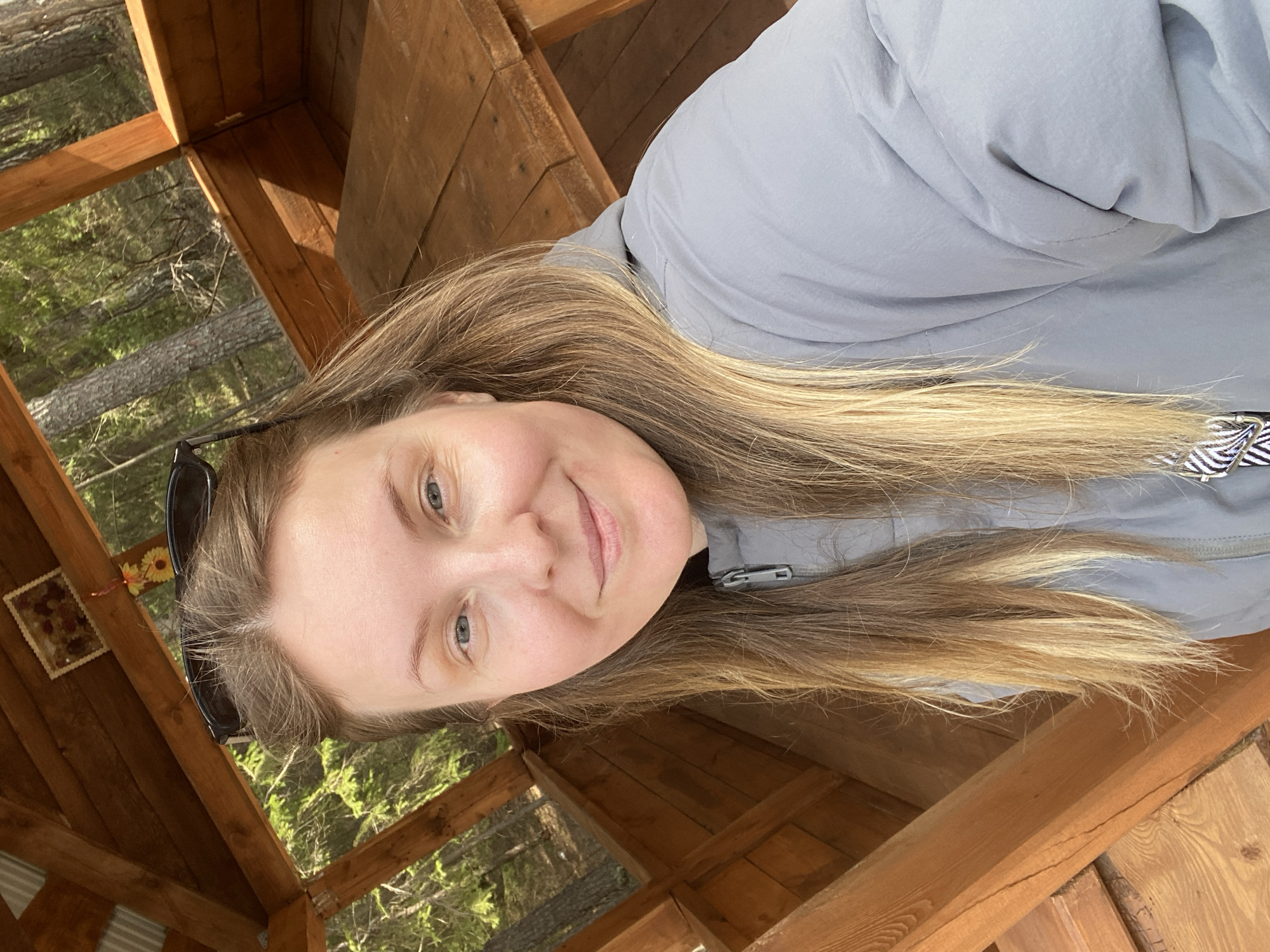 The original image, rotated ninety degrees clockwise, captures a woman smiling softly while taking a selfie. She has fair skin, long light brown and blonde hair, and blue-grey eyes. Sunglasses are perched on her head, and she wears a light grey jacket with a visible zipper. She appears relaxed and content.
The setting is a rustic wooden structure, likely a gazebo or a viewing shelter, with visible beams and planks. Through the openings of the structure, vibrant green trees and foliage are visible, indicating an outdoor, natural environment, possibly a forest or park. A small decorative frame, possibly containing dried flowers or potpourri, hangs on one of the wooden posts. The scene is brightly lit by natural daylight, suggesting clear weather during the day. This peaceful moment is captured in Noviy, Russia. No discernible text is visible.