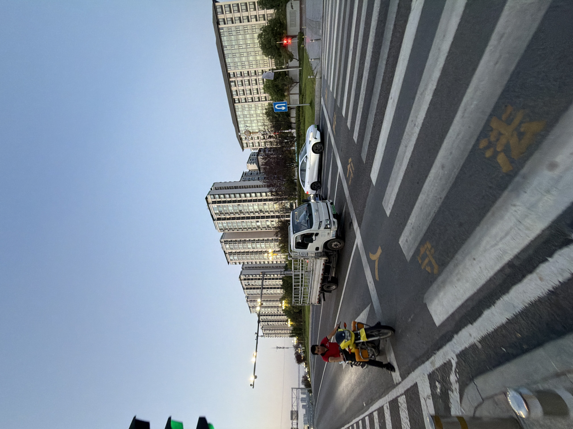 A wide-angle, eye-level shot captures a street scene in the city of Jiangjin, China. The sky is a clear, pale blue, transitioning to a lighter hue near the horizon. In the foreground, a white truck with a silver bed is stopped at a crosswalk, its headlights on. To the left, a man in a red shirt rides a yellow motorcycle, also positioned at the crosswalk. He is looking forward, and his motorcycle has two yellow saddlebags on the rear. The crosswalk itself is marked with wide white stripes. Yellow road markings, including arrows and what appears to be a pedestrian symbol, are visible on the asphalt.
In the middle ground, a white sedan is stopped at the crosswalk, to the right of the truck. A blue sign with a white U-turn arrow is posted on a green grassy area behind the sedan. Further back, a series of tall, modern residential buildings with numerous windows line the horizon, interspersed with trees. Traffic lights are visible on a long overhead arm extending across the road, with what appear to be cameras attached. The overall lighting suggests it is either early morning or late evening.