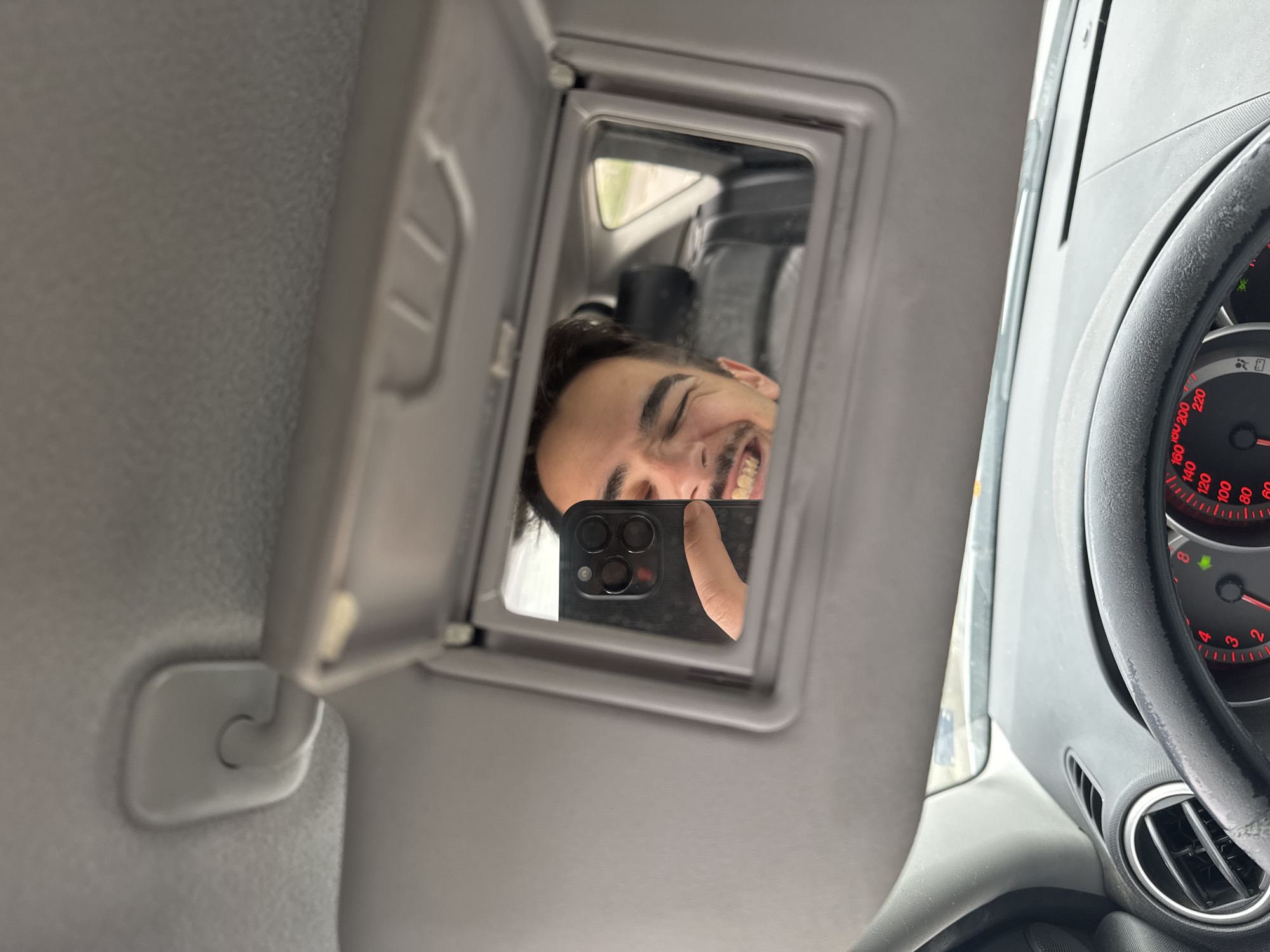 A person is taking a selfie in a car using the mirror on the sun visor. The person is smiling and holding a black smartphone with multiple camera lenses. The dashboard, steering wheel, and speedometer are visible in the background. It's impossible to determine the location in Sofia, Bulgaria, based on this image alone.