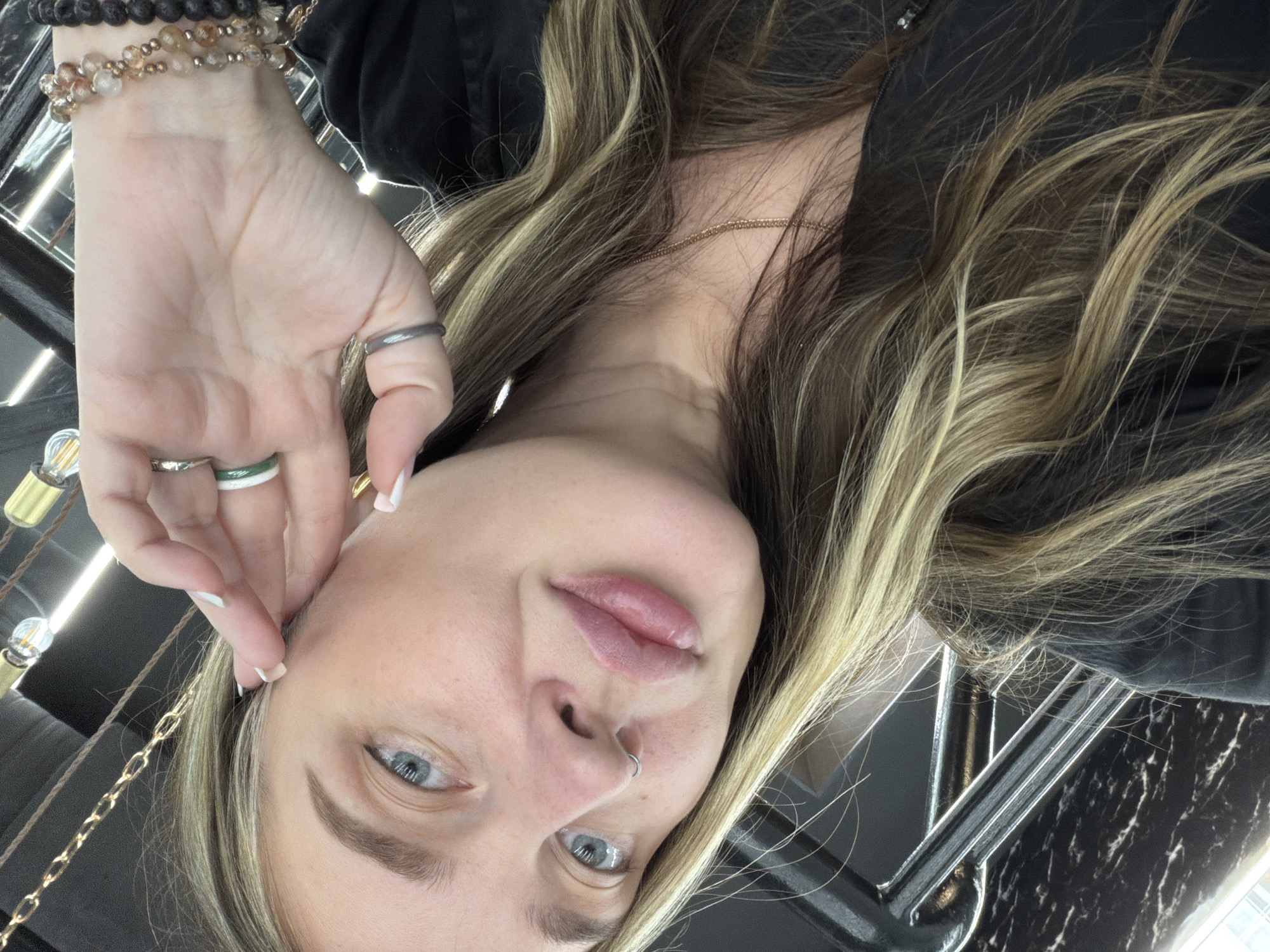 The image is an inverted close-up selfie of a person, likely taken indoors. The subject has fair skin, light blue eyes, and long, wavy hair featuring blonde highlights over darker roots. A subtle silver hoop nose ring adorns their left nostril, and their lips are slightly parted. They are wearing a dark top, possibly black.
The person's left hand is raised towards their face. Their wrist is decorated with multiple beaded bracelets, including darker beads and lighter, translucent ones. Several rings are worn on their fingers: a thin dark band on the index finger, a silver-toned band on the middle finger, and a distinctive white and green striped ring on the ring finger. Their nails are neatly manicured with white tips. A thin gold-colored chain necklace is visible around their neck, and a delicate gold chain earring or ear cuff is near their earlobe.
The background suggests a contemporary indoor environment. Modern light fixtures or metallic architectural elements are reflected prominently. To the right, a dark surface with a white or grey marble-like pattern is visible, possibly a table or counter. The lighting appears bright and artificial. The person's gaze is direct, and their expression is neutral and relaxed. There is no visible text within the image. The location context provided is Moscow, Russia.