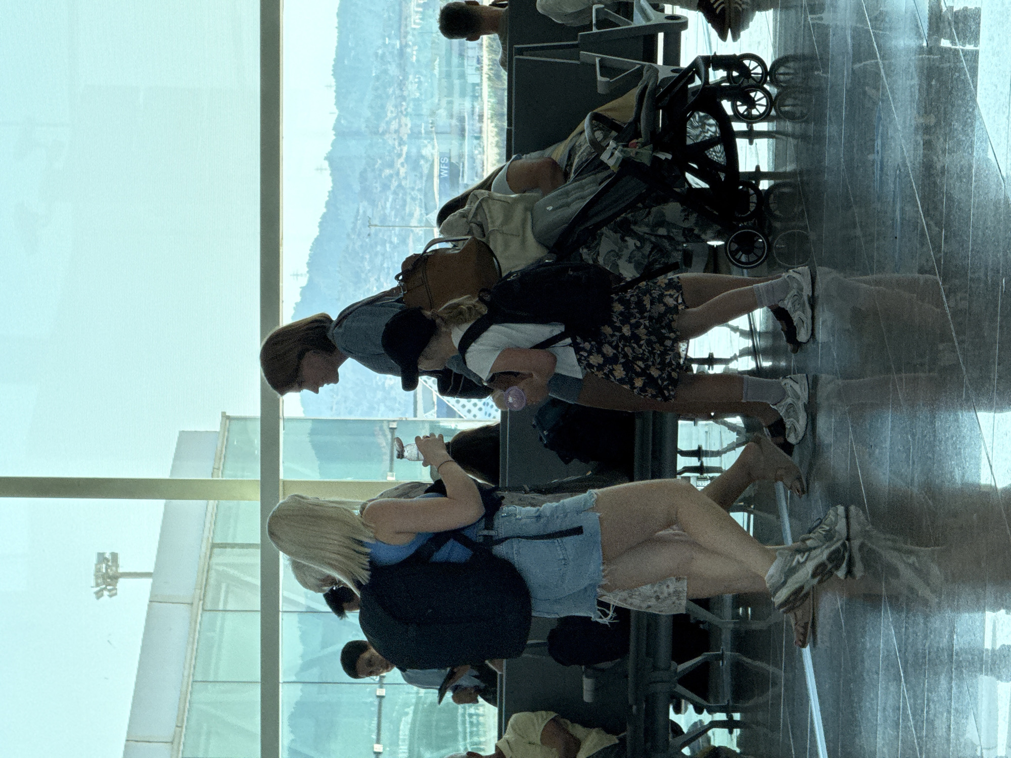 The image shows several people sitting and standing in what appears to be an airport terminal. A stroller is visible, suggesting travel with a child or for convenience. The setting is indoors, with large windows looking out onto a landscape with buildings and hills, possibly indicating a view of the city and its surroundings. The lighting suggests it is daytime. The people are dressed casually, some with backpacks and bags, indicating they are travelers. One person is wearing a blue top and denim shorts, another a floral print skirt, and another a dark hoodie. The floor is reflective, creating mirrored images of the people and objects. The overall mood seems relaxed, as people are waiting. There is no visible text in the image. The location context provided is el Prat de Llobregat, Spain, which is known for being the location of Barcelona–El Prat Airport.