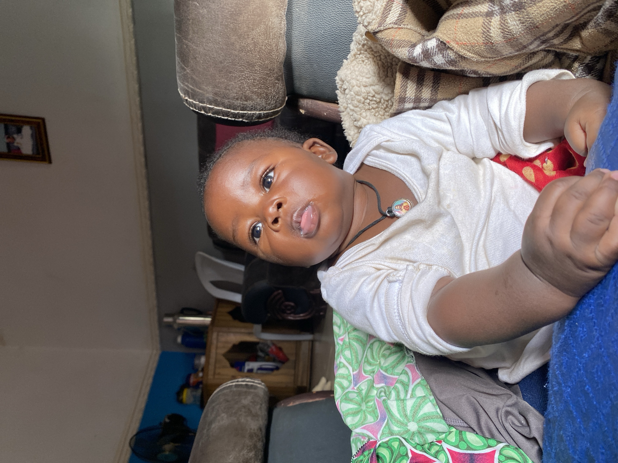 An indoor, portrait-oriented image features a young infant lying down, looking directly at the camera. The baby has dark skin, wide dark eyes, and a slightly open mouth with a tongue peeking out. They are dressed in a long-sleeved white top and wear a black string necklace with a small, circular pendant displaying a colorful, cartoon-like character.
The infant is surrounded by various household textiles and furniture. To the right, a plaid blanket with brown, tan, and white patterns is visible, alongside a cream-colored fluffy blanket. Below the baby, there's a vibrant fabric with green, white, and red leaf-like patterns, and a blue fabric. Parts of what appears to be a brown leather-like sofa or chair are present in the upper and lower left corners, suggesting a comfortable, domestic environment. In the softly focused background, a light-colored wall with a small framed picture can be seen, along with hints of other furniture.
The scene captures a calm, everyday moment of the baby at home, likely in Vom, Nigeria. The lighting suggests an indoor daytime setting. There is no visible text in the image.