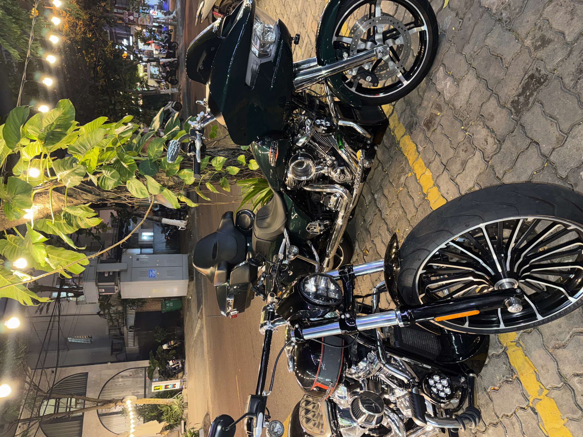 Two motorcycles, one green and one black, are parked on a paved street near a curb marked with yellow paint. The green motorcycle is positioned further back in the frame, partially obscured by a tree with large green leaves. The black motorcycle is closer and more prominent. The scene is in Đà Nẵng, Vietnam, illuminated by overhead string lights. A building is in the background. Other vehicles are visible in the distance. No people are visible in the scene.