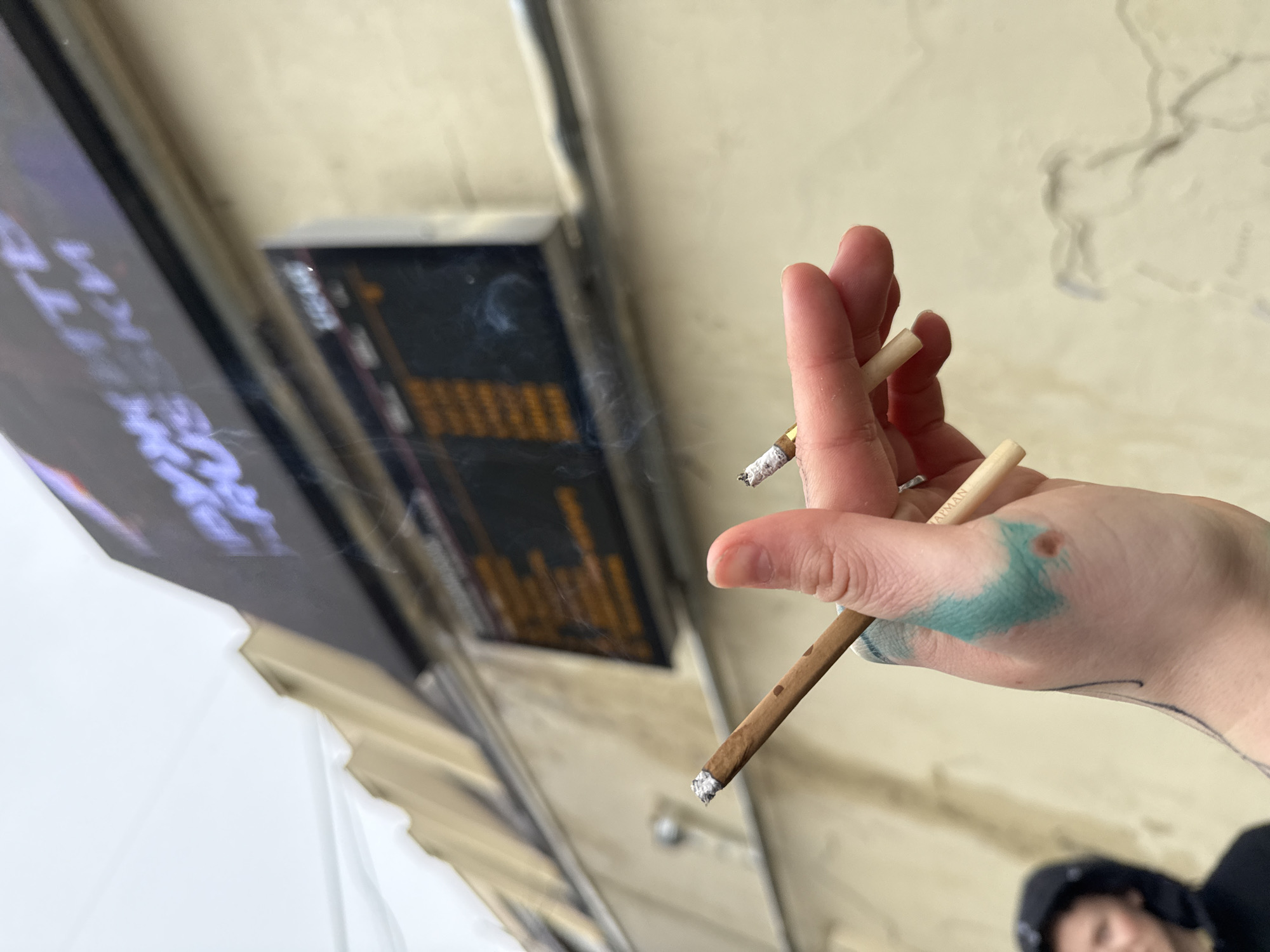 A hand holds a lit cigarette and a thin, unlit wooden rod. The lit cigarette is held between the thumb and forefinger, while the wooden rod is held between the middle and ring fingers. Smoke is rising from the lit cigarette. The hand has a teal-colored tattoo on the back. A blurry image of a person wearing a hooded garment is visible in the lower right corner of the image. In the background, there is a digital sign with blurred content. The image appears to be taken outdoors, possibly near an urban transit hub, in the city of Unknown, Unknown.