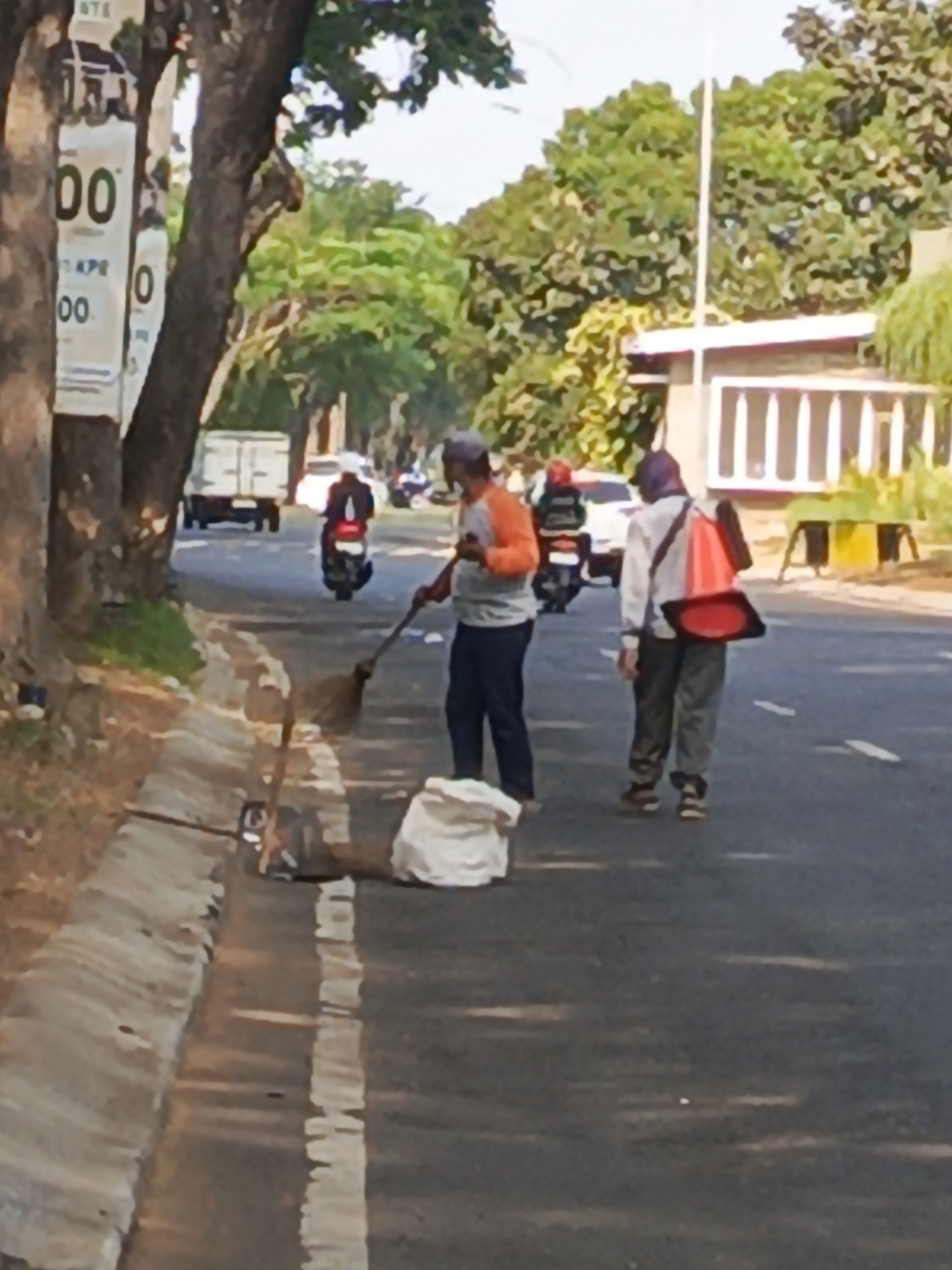 The image shows a street scene in Calukan RT 1 RW 5 Keboansikep, Indonesia. Two men are present. One man, wearing an orange shirt and dark pants, is sweeping the roadside with a broom. A white garbage bag is situated next to him. The second man, wearing a white shirt, dark pants, and a purple cap, walks away from the viewer, carrying a red traffic cone on his back, secured by a strap. A motorcycle is visible in the background, with a rider wearing a white helmet and black jacket. A white truck and another motorcycle are also in the background on the road. Trees line the left side of the road, with some signage attached to one of the trees. The environment appears to be a semi-urban or suburban area with a paved road and a grassy verge.
