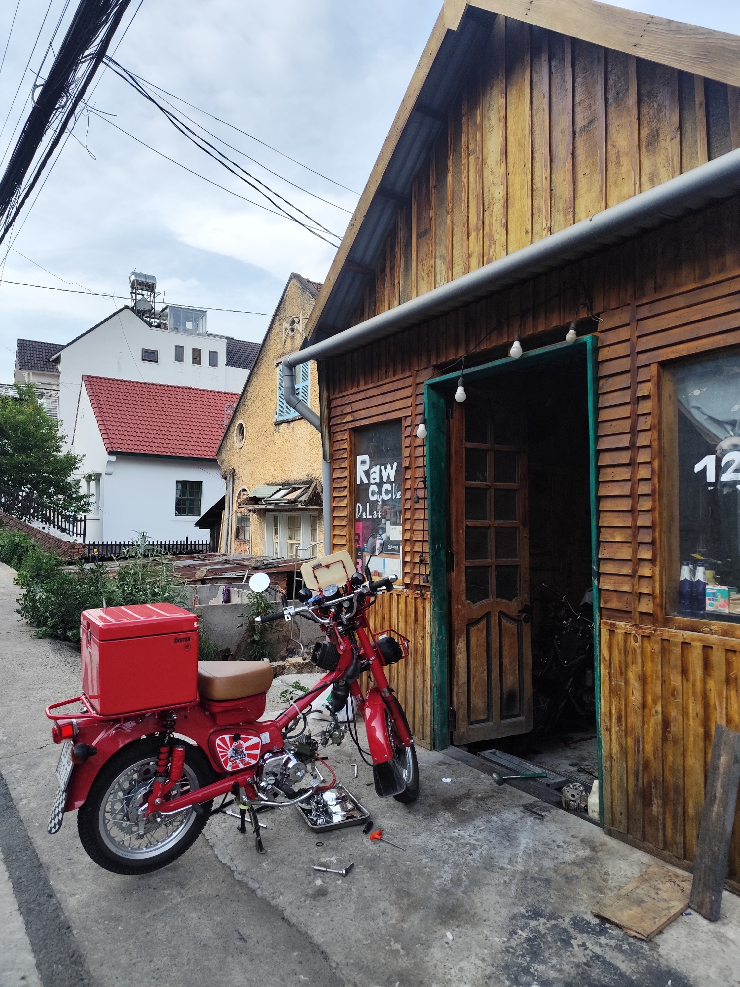 In Cam Ly - Đà Lạt, Vietnam, a red motorcycle with a red delivery box is parked on the side of a street in front of a rustic wooden building. The motorcycle is partly disassembled, with tools and parts laid out on a tray nearby. The building features a green-trimmed door that is open, revealing a dimly lit interior. A sign is visible on the window to the left of the door, partially obscured but reading "Raw C. Cycle...Da Lat". Power lines are overhead, with houses and vegetation visible in the background. The sky is overcast.