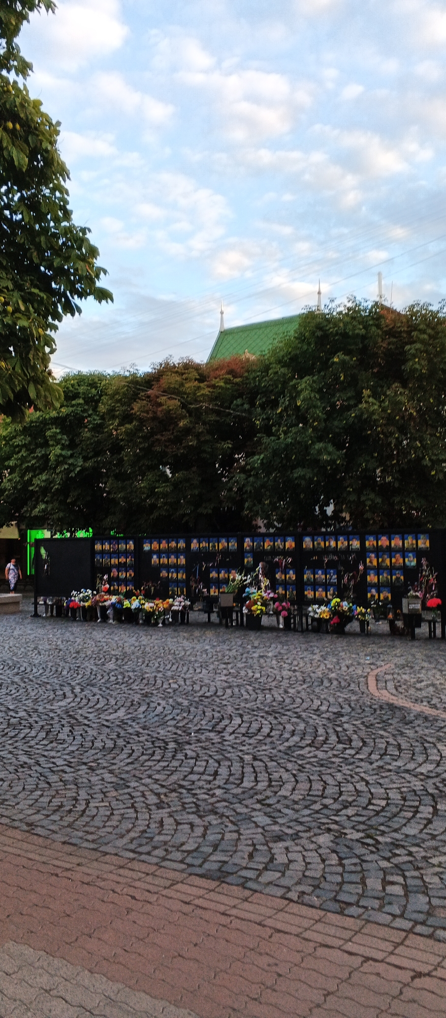 In Mukachevo, Ukraine, a memorial display features a large black wall adorned with numerous framed portraits, each likely of an individual. In front of the wall, an arrangement of buckets filled with colorful flowers, predominantly yellow, red, and white, creates a somber tribute. The scene is set on a cobblestone plaza, with a woman in a patterned dress walking on the left side of the frame. Overhead, a lush green tree provides shade, and a building with a distinctive green roof and white spires is visible behind the trees. The sky above is a mix of blue and white clouds.