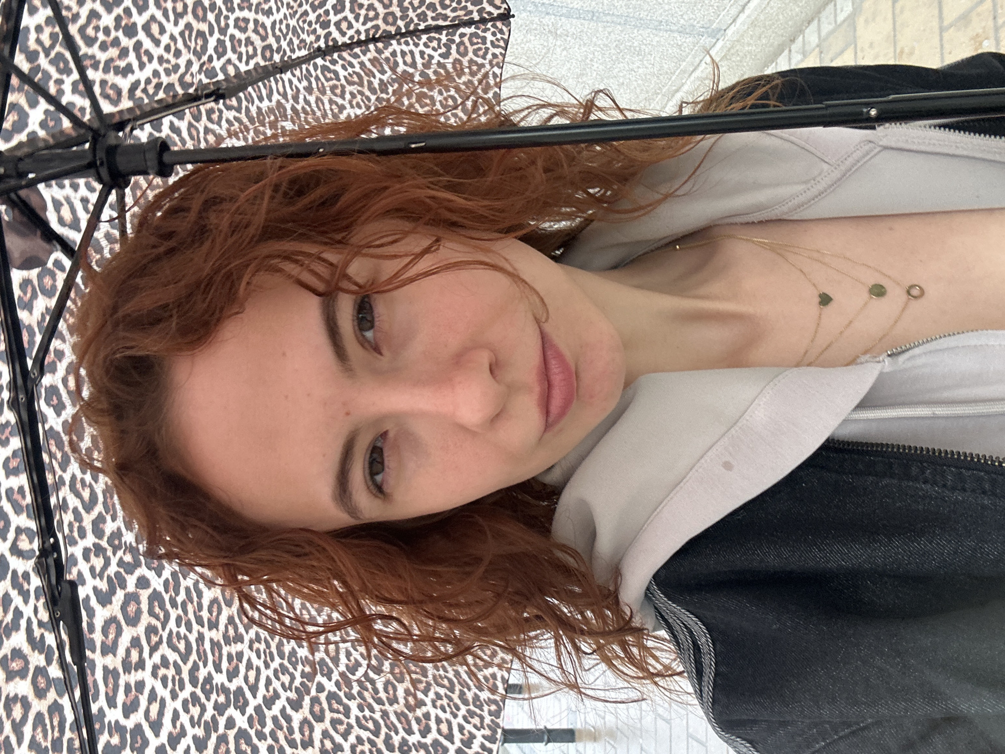 A selfie taken by a woman with red, curly hair under a leopard-print umbrella. The woman is fair-skinned with light-colored eyes. She is wearing a light-colored hoodie with a dark denim jacket over it and a multi-strand gold necklace with small charms. The background appears to be a light-colored sidewalk or paving. The location is Novi Sad, Serbia.