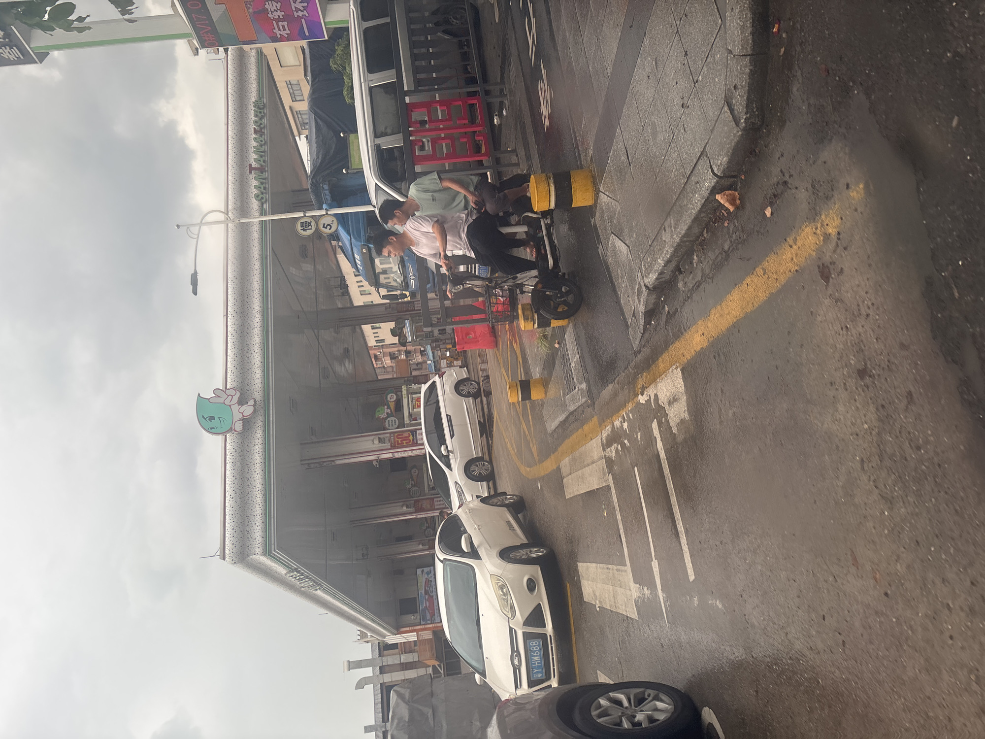 In Nanhai District, China, a gasoline station with a white canopy and the name "SANTANGSHI" emblazoned on it is visible. The sky is overcast. Two men are riding an electric scooter with a basket on the front, heading towards the station. A white Ford Focus, with license plate "BY HW688," and a white minivan are parked in front of the station. Yellow and black bollards are placed on the wet asphalt. Traffic markings, including pedestrian crossing lines, are visible on the road.