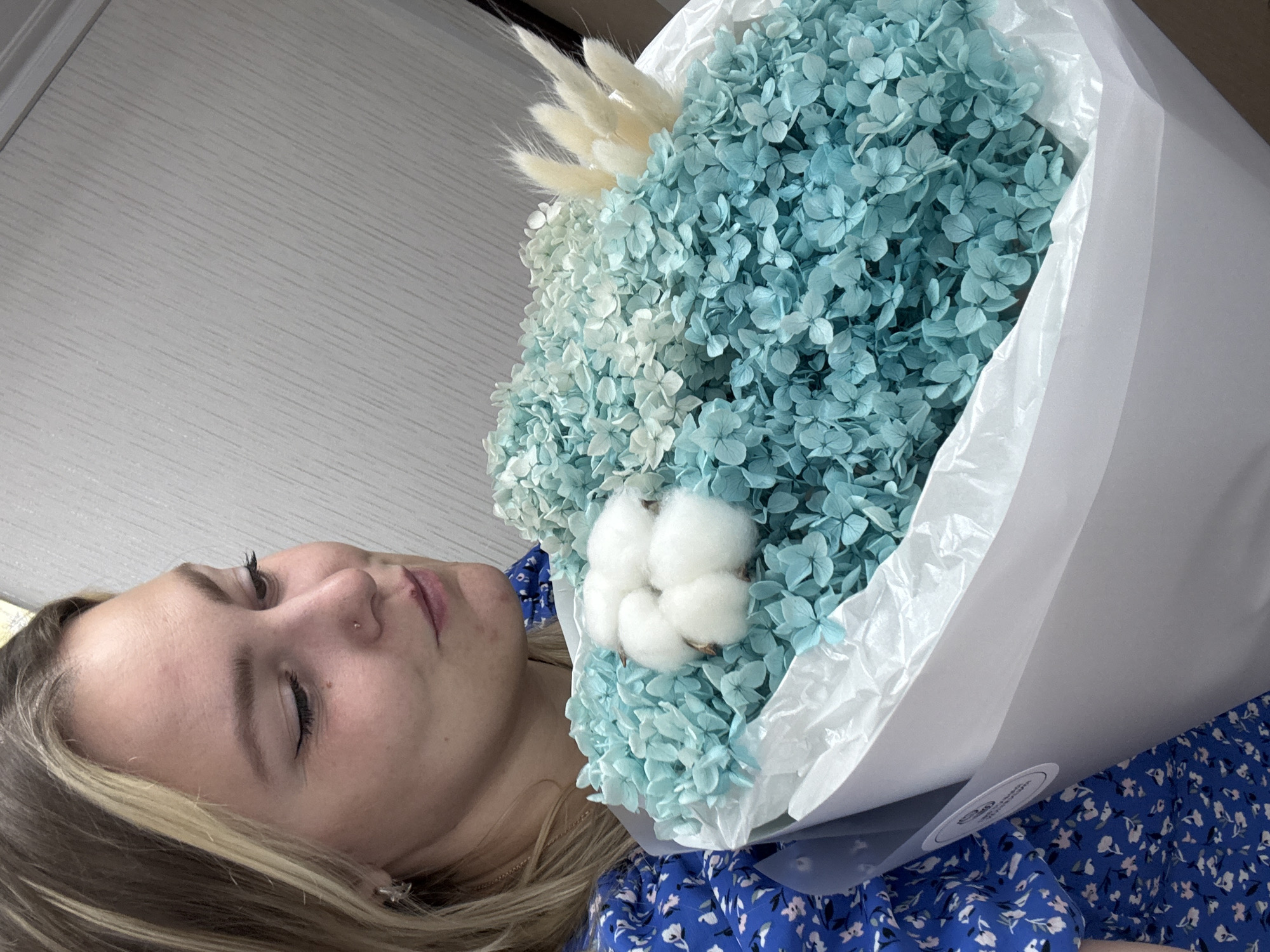 A young woman with blonde hair, wearing a blue floral dress, holds a bouquet of dried blue hydrangeas and cotton balls. The bouquet is wrapped in white paper. The woman is looking down at the flowers with a gentle smile. The background appears to be a light-colored wall with a subtle striped pattern. The location is identified as Noviy, Russia.