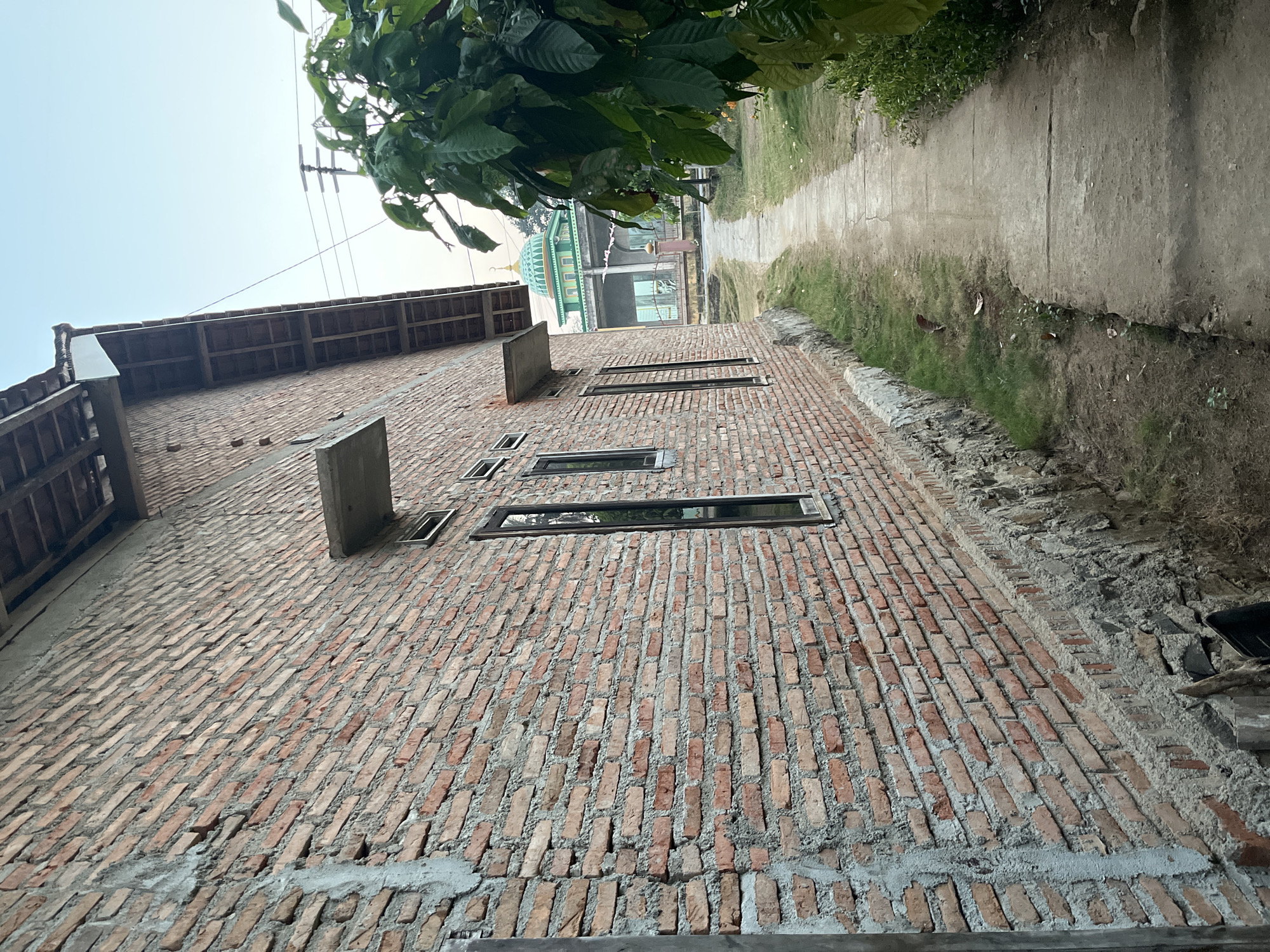 The image shows a narrow concrete pathway leading towards a green-domed mosque in the distance. On the left side of the pathway is a brick building with a rough concrete foundation. The building has several windows with dark frames. On the right side of the pathway, lush green foliage is visible. The sky is clear and light. The scene appears to be in Kebumen, Indonesia.