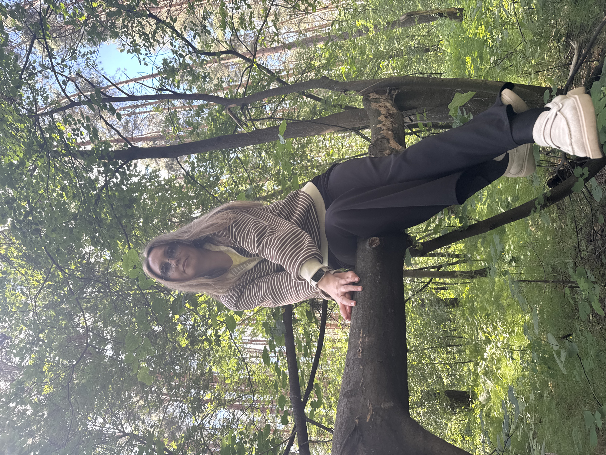A woman with long blonde hair and glasses is sitting on a fallen tree trunk in a forest. She is wearing a striped sweater over a yellow top and black pants. Her feet are clad in white slip-on shoes. The background is filled with green trees and foliage. The lighting suggests it is daytime. The location is identified as Chaikovsky, Russia.