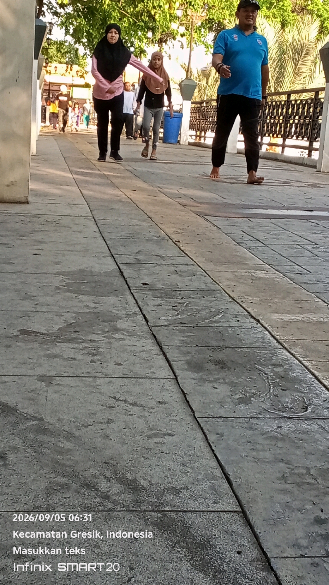 The image shows a street scene in Gresik, Indonesia, on September 5, 2026, at 06:31. A person wearing a black hijab and a pink shirt walks on a paved walkway, with their right arm extended. To their left and slightly behind, a child in a brown hijab and grey leggings walks, also on the walkway. Further to the right, a man wearing a blue t-shirt and black pants, with a cap, is walking barefoot on the same walkway. In the background, there are trees, a metal railing, and a blue trash can. The walkway is made of concrete slabs. The text "Masukkan teks" is also visible, which translates to "Enter text."