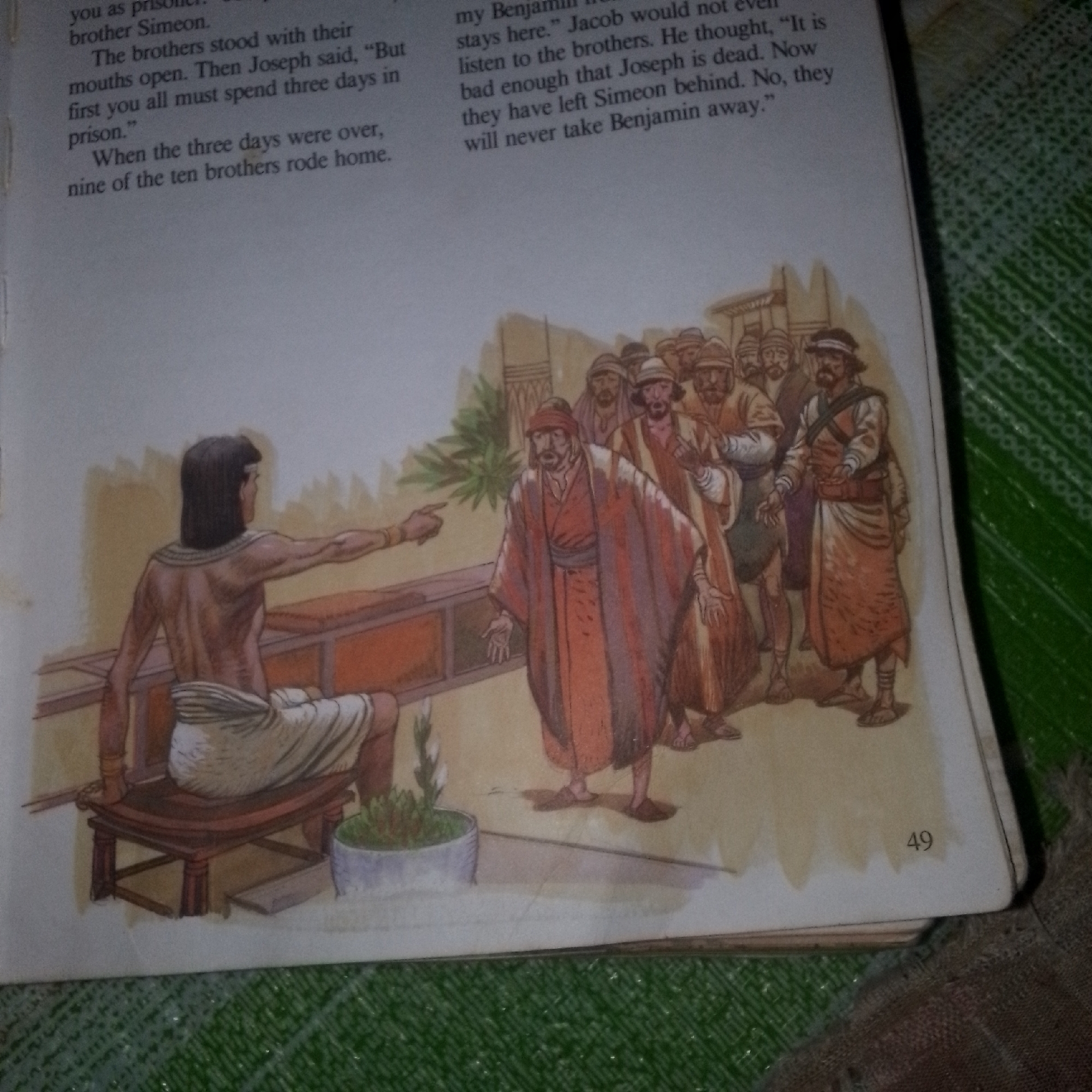 In an indoor scene, a man is seated on a stool, facing away from the viewer and gesturing to a group of men. The seated man has dark hair and wears a white cloth around his waist and a gold necklace. The group of men are standing and appear to be listening to the seated man. One man in the group, wearing an orange robe, is gesturing with his hands. The scene is depicted in a watercolor style. The page has text on the left side and the number "49" is visible in the lower right corner. The background is a pale yellow, suggesting an interior setting.