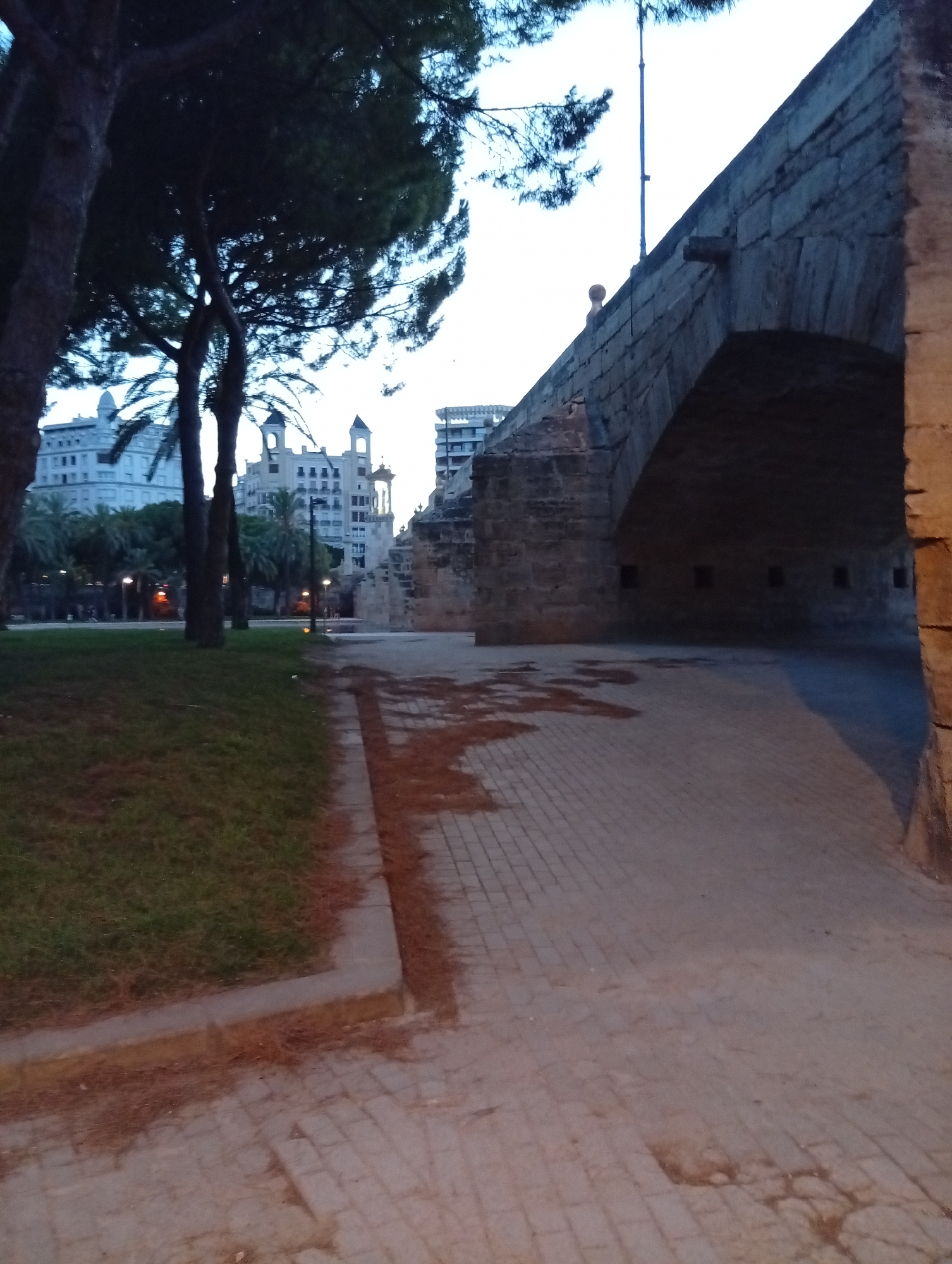 The image shows a view of the Towers of Serranos in Valencia, Spain, at dusk. In the foreground, a paved walkway runs alongside a grassy area. A large stone archway, part of the Towers of Serranos, dominates the right side of the image. The walkway leads under the archway into a darker area where several small rectangular openings are visible in the stone structure. In the background, across a street or plaza, are several buildings, including white buildings with ornate features and taller, modern structures. Palm trees are visible between the walkway and the buildings. The sky is a pale, dusky hue.