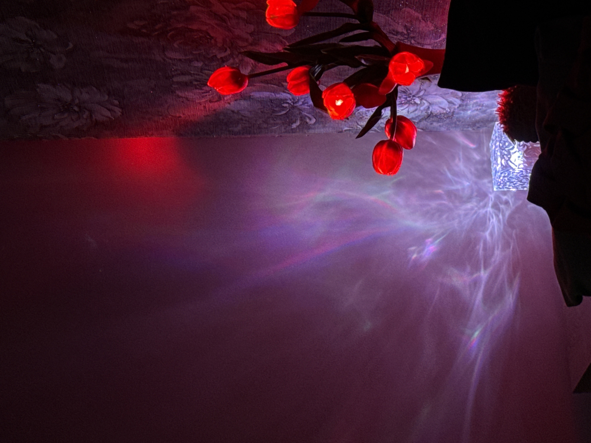 Here's a description of the image:
The image presents a low-angle view within a room. A decorative light fixture hangs from the ceiling, featuring multiple red, tulip-shaped lights. The light reflects off the ceiling, which has a patterned wallpaper. Below the fixture, on the wall, are patterns of light in hues of purple, blue, and white. The light is angled towards the wall. A portion of a person is seen to the right of the image. Part of an illuminated object is seen between the fixture and the person.