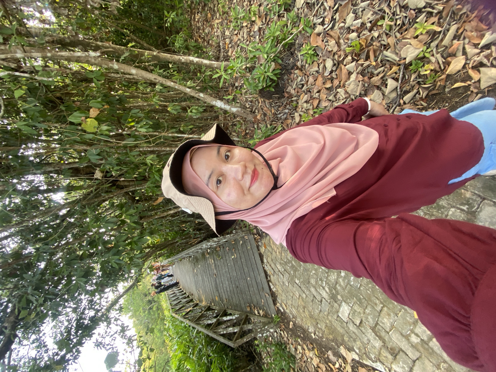 In the city of Sungai Pitung, Indonesia, a woman wearing a pink hijab and a maroon top with a hat is taking a selfie. She is standing on a brick pathway that leads to a wooden bridge. The bridge is lined with trees and lush greenery. A group of people are walking on the bridge in the distance. The ground is covered with fallen leaves, indicating an outdoor, natural environment.