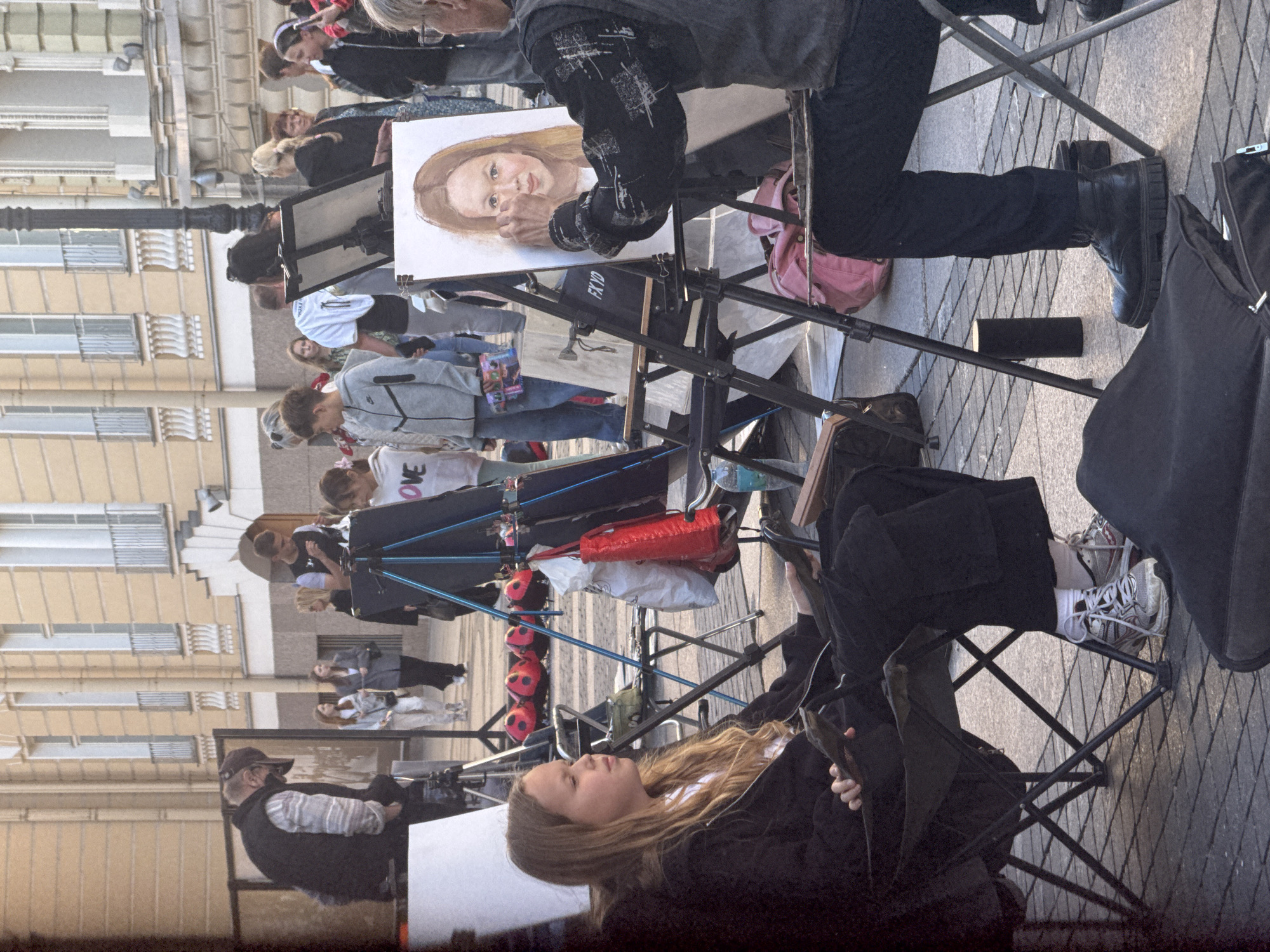 The image depicts an outdoor art session in St. Petersburg, Russia, likely on Palace Square, given the architectural style of the buildings in the background. A young girl with blonde hair is seated in a black folding chair, facing to the right, wearing a black zip-up hoodie and black pants. She is holding what appears to be a book or tablet in her lap. In front of her, an artist, partially visible from the side and back, is working on a portrait of the girl on an easel. The artist is seated in a chair and wearing dark clothing. The portrait on the easel shows a young girl with blonde hair and fair skin.
Several other people are present in the background, milling about in what seems to be a public square. One person is seen walking with another. There are also several red, inflatable objects, shaped like ladybugs, scattered on the ground. A white bag and a red bag are hanging from a tripod structure to the right of the girl. The ground is paved with cobblestones. The buildings in the background are light-colored, with multiple windows and architectural detailing, consistent with St. Petersburg's classical architecture. The overall scene suggests a casual, daytime artistic activity in a public space.