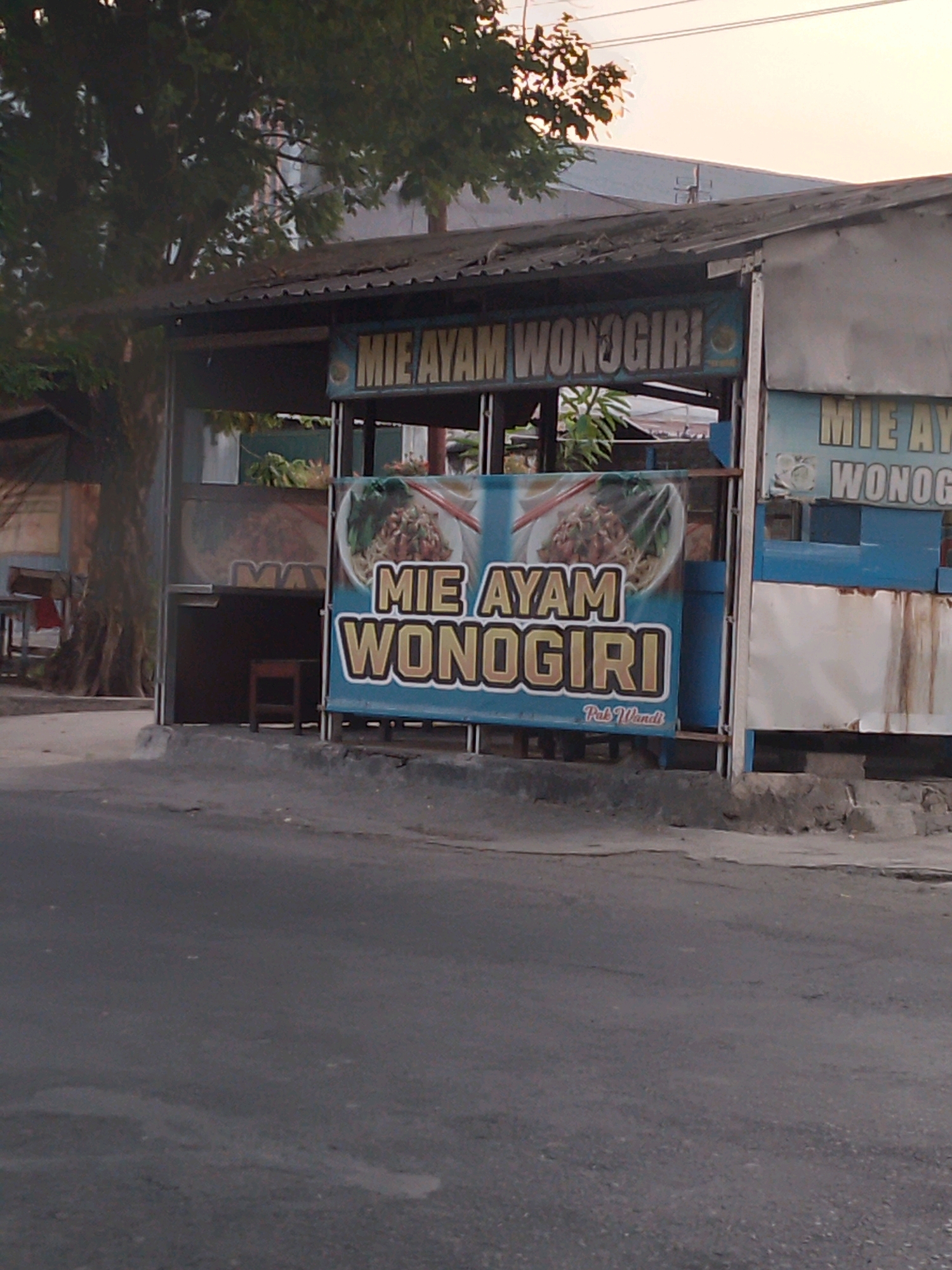 The image shows a street-level view of a food stall in Kertosono, Indonesia, likely in front of a building. The stall has a corrugated metal roof supported by poles. A prominent banner on the front of the stall displays the text "MIE AYAM WONOGIRI" in large, bold letters, with an image of a noodle dish above. The stall appears to be open for business, with a counter and some seating visible inside. There is a large tree to the left of the stall, and the street in front is paved. The overall lighting suggests it is either early morning or late afternoon.