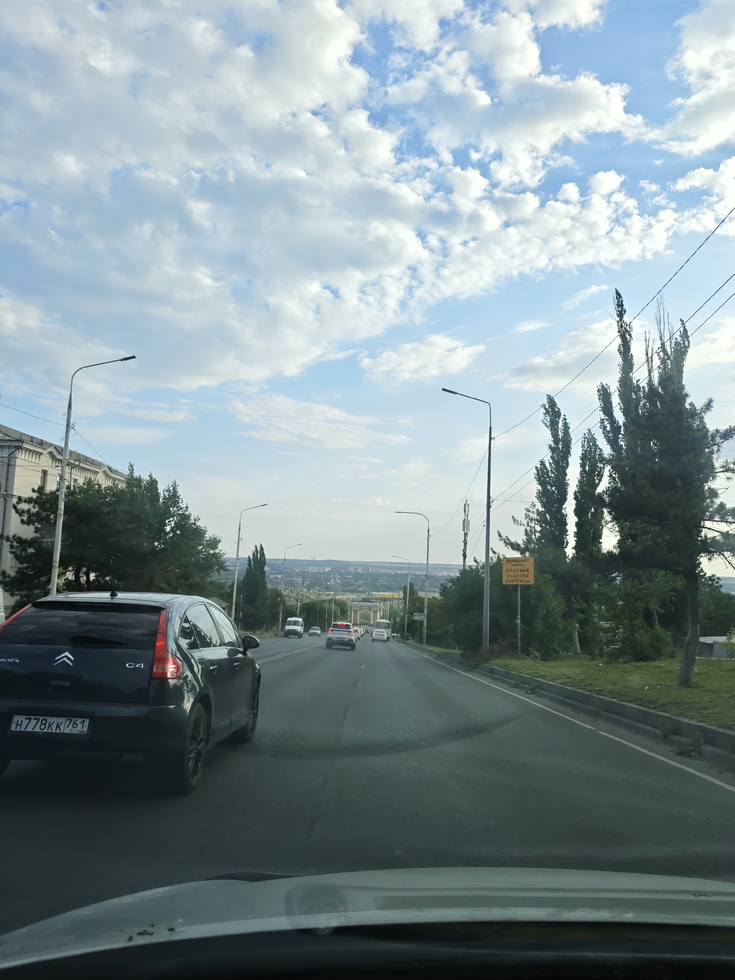The image shows a view from a vehicle driving on a road in Novocherkassk, Russia. The sky is partly cloudy with a mix of blue sky and white cumulus clouds. On the left side of the road is a building with arched windows and trees in front of it. The road itself has multiple lanes of traffic. Several cars are visible, including a dark-colored Citroën C4 in the foreground on the left, a white van, and other vehicles further down the road. There are streetlights and power lines along the side of the road. A yellow sign with Russian text warning of danger is visible on the right side of the road, near tall, slender trees. The road appears to be an urban or suburban thoroughfare, with greenery and distant buildings indicating the city of Novocherkassk.