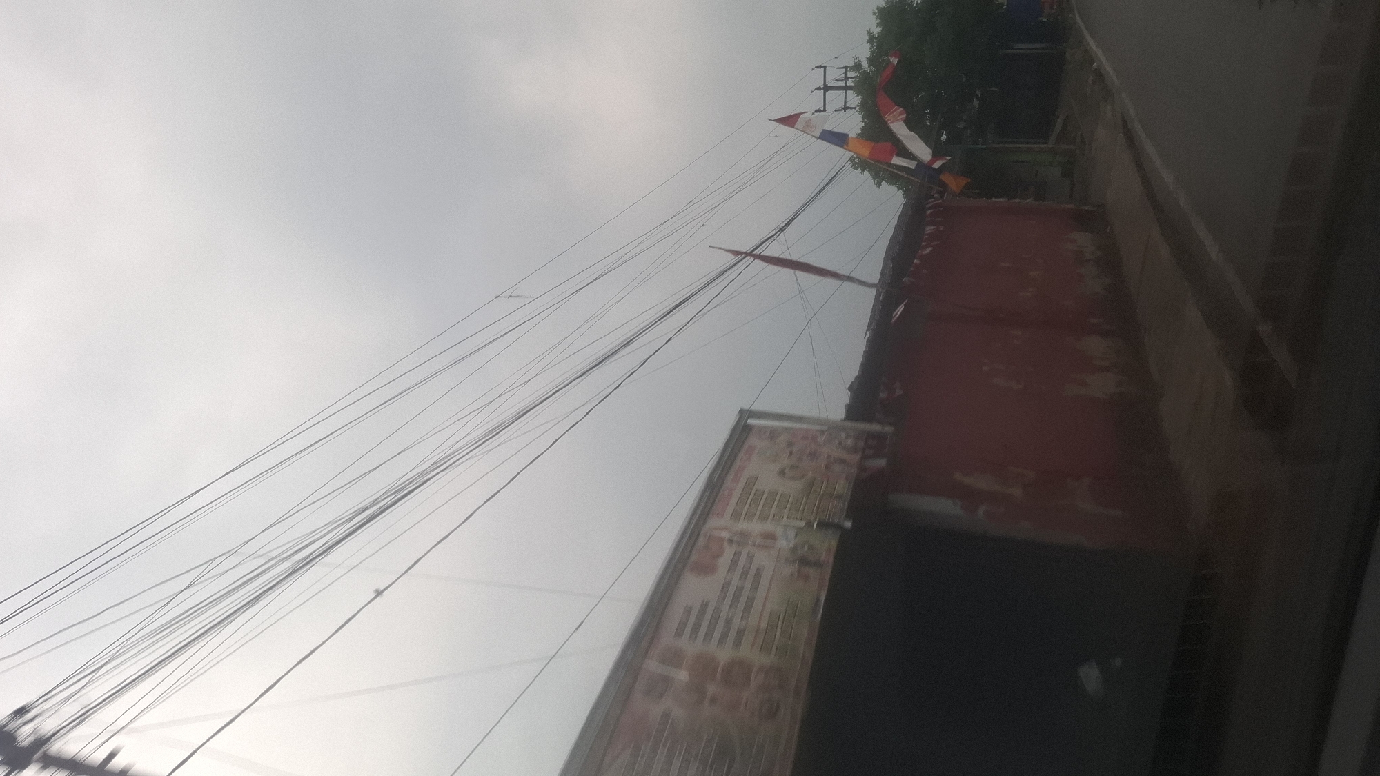 The image shows a street scene in Sirnagalih, Indonesia, under an overcast sky. A red building with a sign is visible on the left, and multiple electrical wires crisscross the sky above. Two Indonesian flags, red and white, are flying from a pole on the right side of the building, accompanied by smaller triangular flags in red and white. A paved road runs alongside the building, and a tree can be seen behind the flags. The overall impression is of a roadside view with a local establishment.