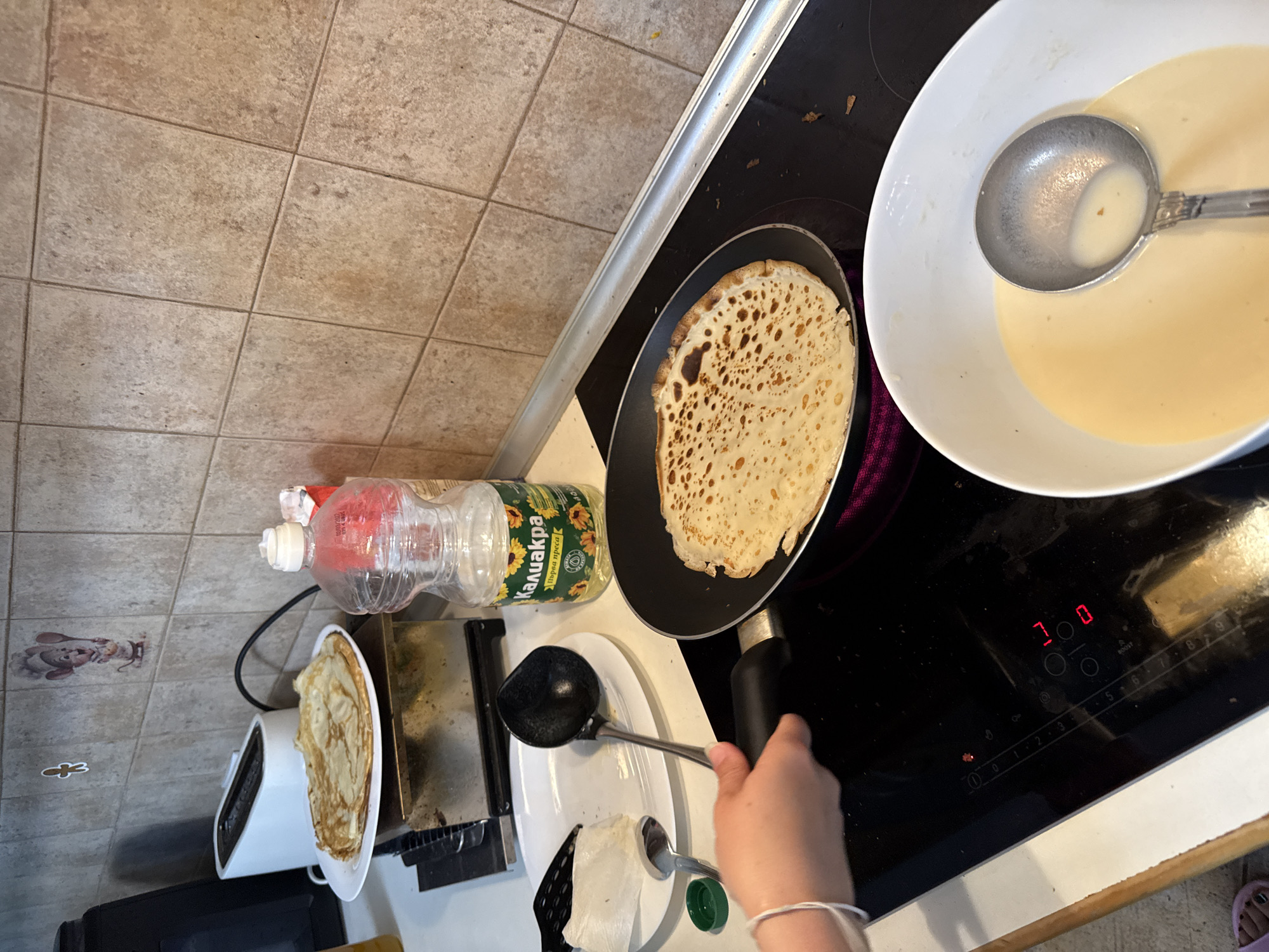 In a kitchen, a person is cooking pancakes. A stack of cooked pancakes sits on a plate next to a toaster. A bottle of vegetable oil is also visible. In the foreground, a hand is holding a ladle over a frying pan with a pancake cooking in it. A white bowl filled with pancake batter is nearby. The stove is electric and displays a red light. The kitchen has tiled walls. The image is taken from a medium angle, showing the top of the stove and countertop.