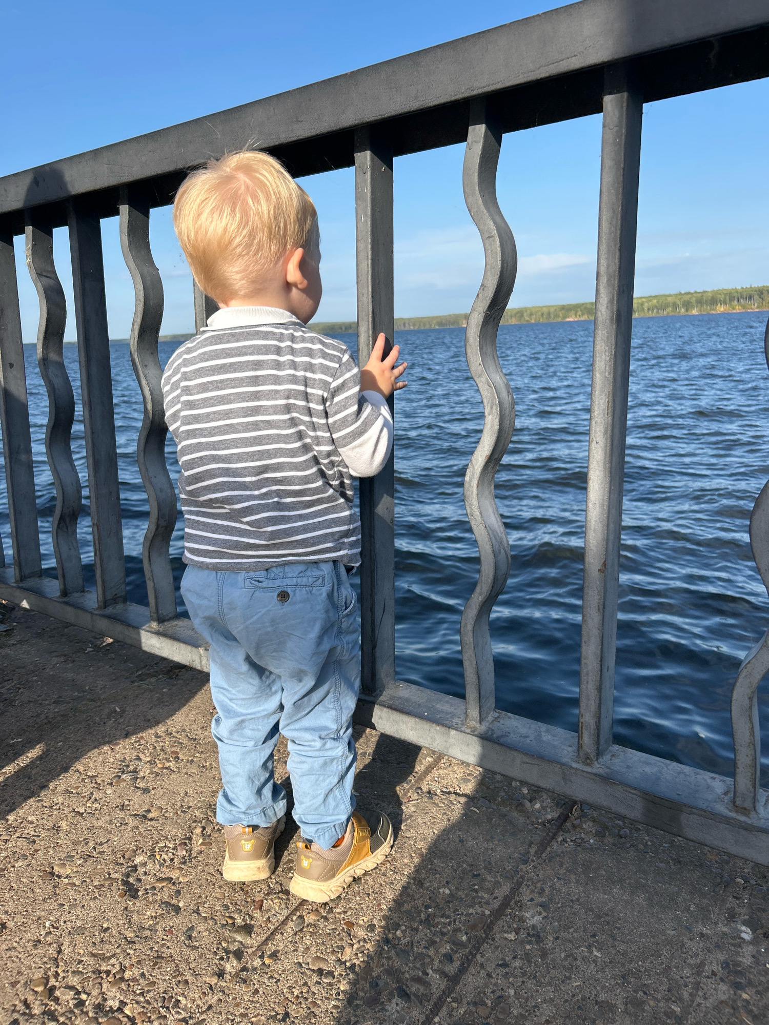 A young boy with blond hair stands facing away from the camera, looking out at a body of water. He is wearing a gray and white striped long-sleeved shirt and light blue pants. He has on tan and gold sneakers. The boy is standing on a concrete surface next to a dark gray metal railing. The railing has decorative wavy bars. Beyond the railing, the water is blue and calm, with a faint tree line visible in the distance under a clear blue sky. The location appears to be a waterfront promenade or park area, possibly in the city of Chaikovsky, Russia, given the context.