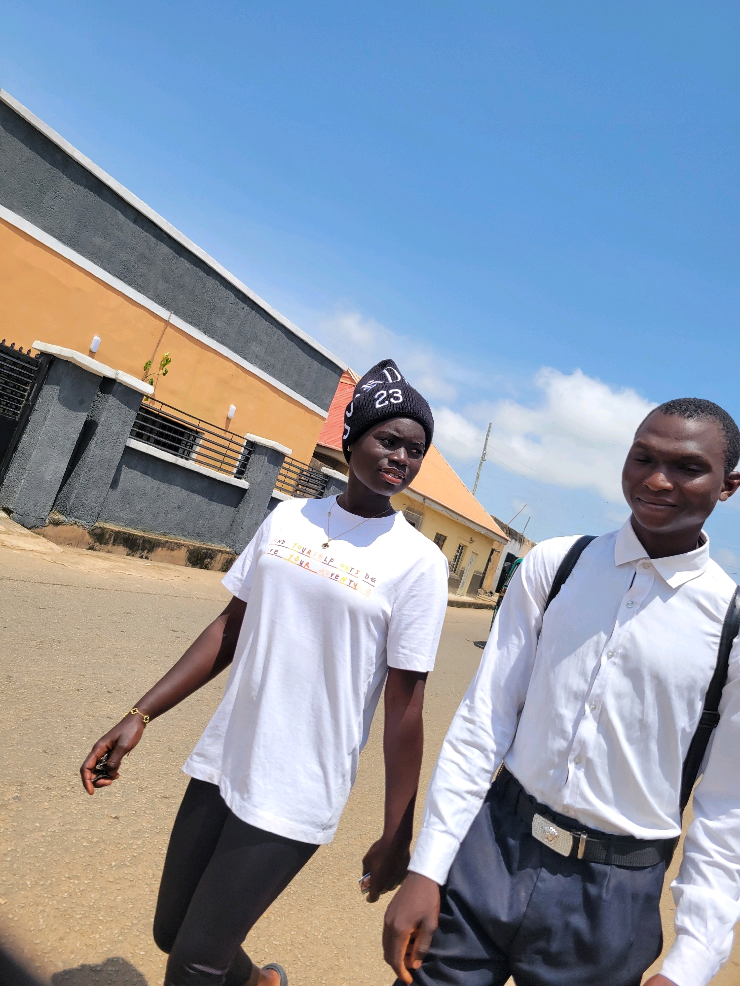 Two young individuals, a female and a male, are captured in a medium shot walking outdoors on a paved street under a bright, clear blue sky with scattered white clouds. The scene appears to be in the city of Jos, Nigeria. The female, on the left, is wearing a black beanie with white lettering that reads "DJ 23", a white t-shirt with colorful text, and black leggings. She has a gold necklace and a bracelet on her left wrist, and she is holding something in her left hand. The male, on the right, is wearing a white button-down shirt, dark trousers, and a black belt with a silver buckle. Suspenders are visible on his shoulders.
Behind them, a building with an orange lower section and a dark grey upper section, separated by a white trim, is partially visible. The building has a fence with black metal bars. Further in the background, more buildings with orange roofs and yellow walls are seen. The overall environment suggests a sunny day in an urban or semi-urban setting. The lighting indicates it is midday.