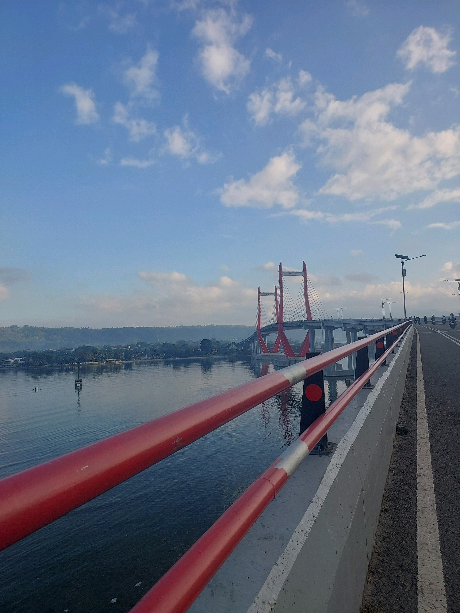This is a landscape photograph of the Merah Putih Bridge in Ambon, Indonesia, taken from the bridge itself. The bridge features a distinctive red and white cable-stayed design with two prominent red towers. The viewpoint is from the right side of the bridge, looking towards the distance where the bridge spans a body of water.
In the foreground, a red railing runs along the edge of the bridge. The railing is made of thick cylindrical pipes and supported by black posts with red circular markers. The railing is in sharp focus, creating a sense of depth.
To the right of the railing is a paved road with white lane markings. Several motorcycles are visible in the distance on the road, suggesting ongoing traffic. A solar-powered street lamp stands tall beside the road.
The bridge extends across a calm body of water. The water reflects the sky and the bridge structure. On the far side of the water, a lush green coastline is visible, dotted with buildings and trees, indicating a populated area. Hills rise beyond the coastline, fading into a hazy blue in the distance.
The sky above is a clear blue, punctuated by scattered white, fluffy clouds. The lighting suggests it is either early morning or late afternoon, with soft, diffused light. The overall atmosphere is serene and picturesque, capturing the architectural marvel of the bridge against the natural beauty of Ambon.
