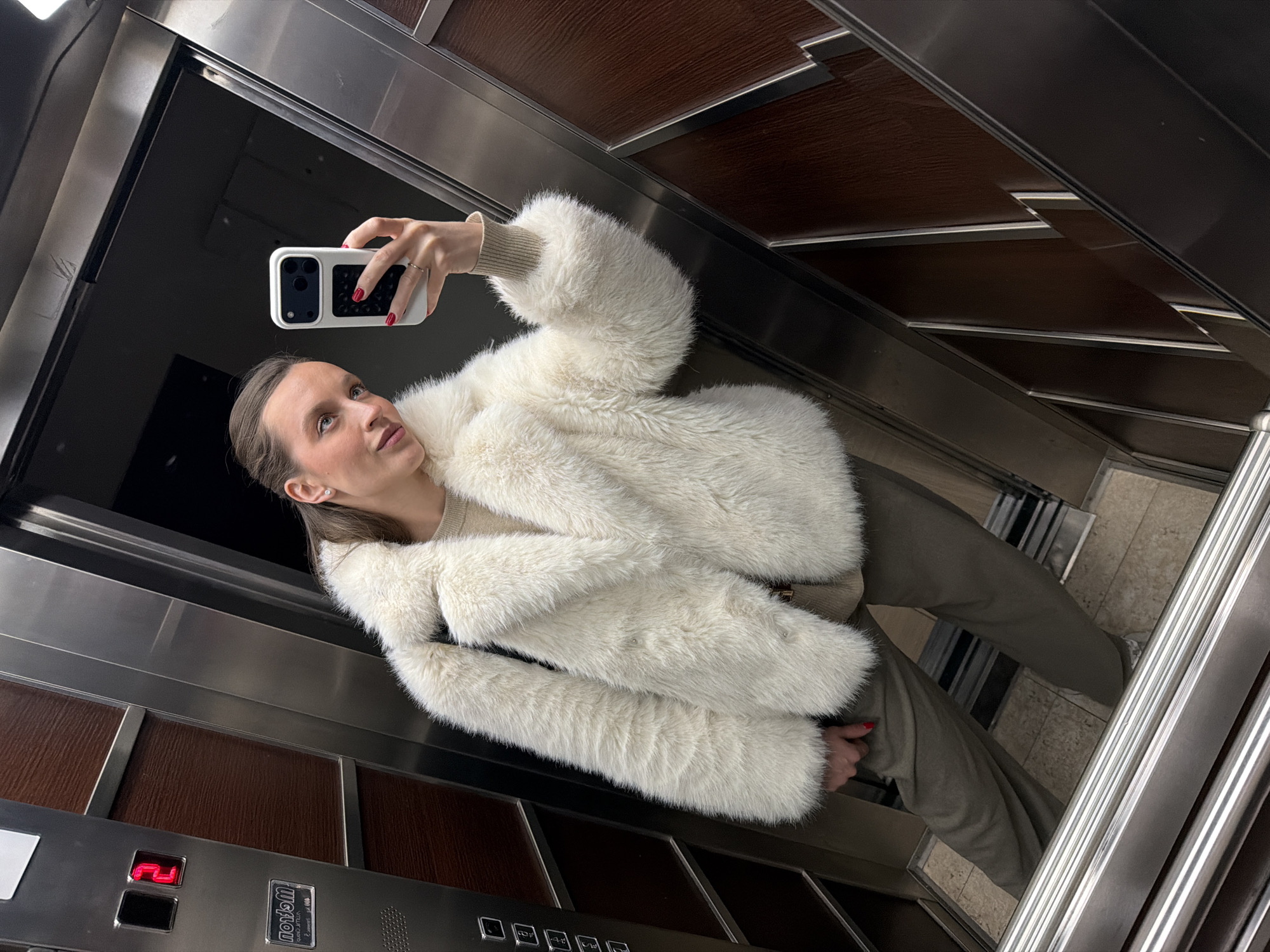 The photograph, taken inside an elevator, depicts a woman holding a white smartphone and capturing a selfie. She is wearing a plush, off-white jacket over a light-colored top and olive-green pants. The elevator's interior is a mix of stainless steel and wood paneling. A floor indicator displays the number "2." Markings indicate the manufacturer is "wotan." While the image does not provide explicit details that confirm the location, this cannot be definitively ascertained without more information.