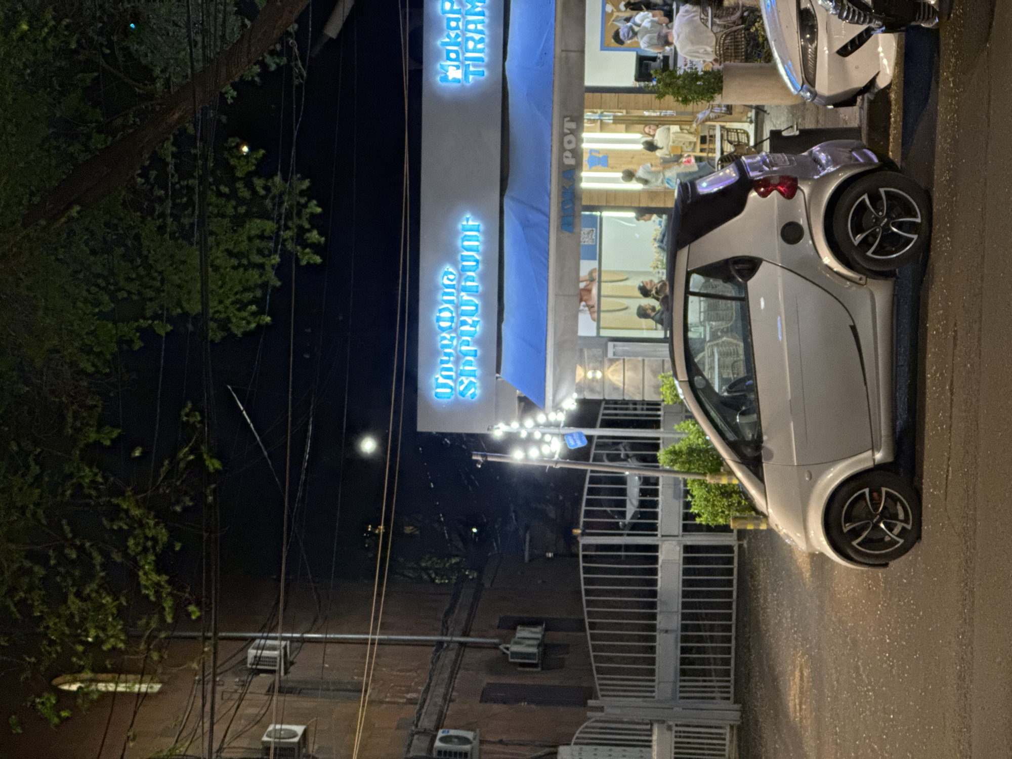 Here is a detailed description of the image:
The image depicts a street scene at night in Yerevan, Armenia. A compact silver car, specifically a Smart car, is parked on the right side of the frame. It appears to be parked near the Moka Pot Cafe, whose illuminated signage in Armenian and English is visible. The cafe has both indoor and outdoor seating areas; people can be seen sitting at tables inside. Adjacent to the Smart car is another parked car.
In the background, there is a gate, utility poles with power lines and electrical equipment attached to the building. There is a tree to the left. The street surface is visible and appears to be wet or damp. The lighting is a mix of streetlights, cafe lighting, and the ambient glow of the city at night.