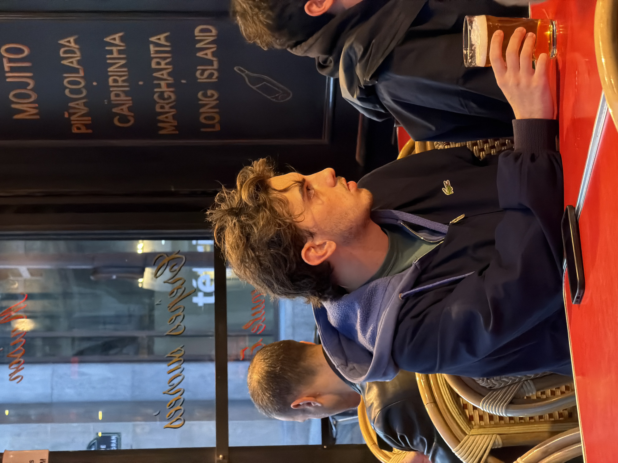 The image captures an indoor scene within what appears to be a restaurant or bar in Paris, France. The foreground is dominated by a man seated at a table. He has dark, curly hair and is wearing a blue jacket with a visible Lacoste logo. He is looking to the left of the frame, with a thoughtful expression. Another person is partially visible next to him. Beside the table, a hand holds a pint of beer. The table is red.
Behind the man, there is a black wall menu listing cocktails, including Mojito, Piña Colada, Caipirinha, Margarita, and Long Island. Further back, a large window offers a glimpse of the outside, where reflections show script text in French. This suggests a typical Parisian establishment with mirrored windows and classic signage. In the reflections, street details are visible, but hard to make out.