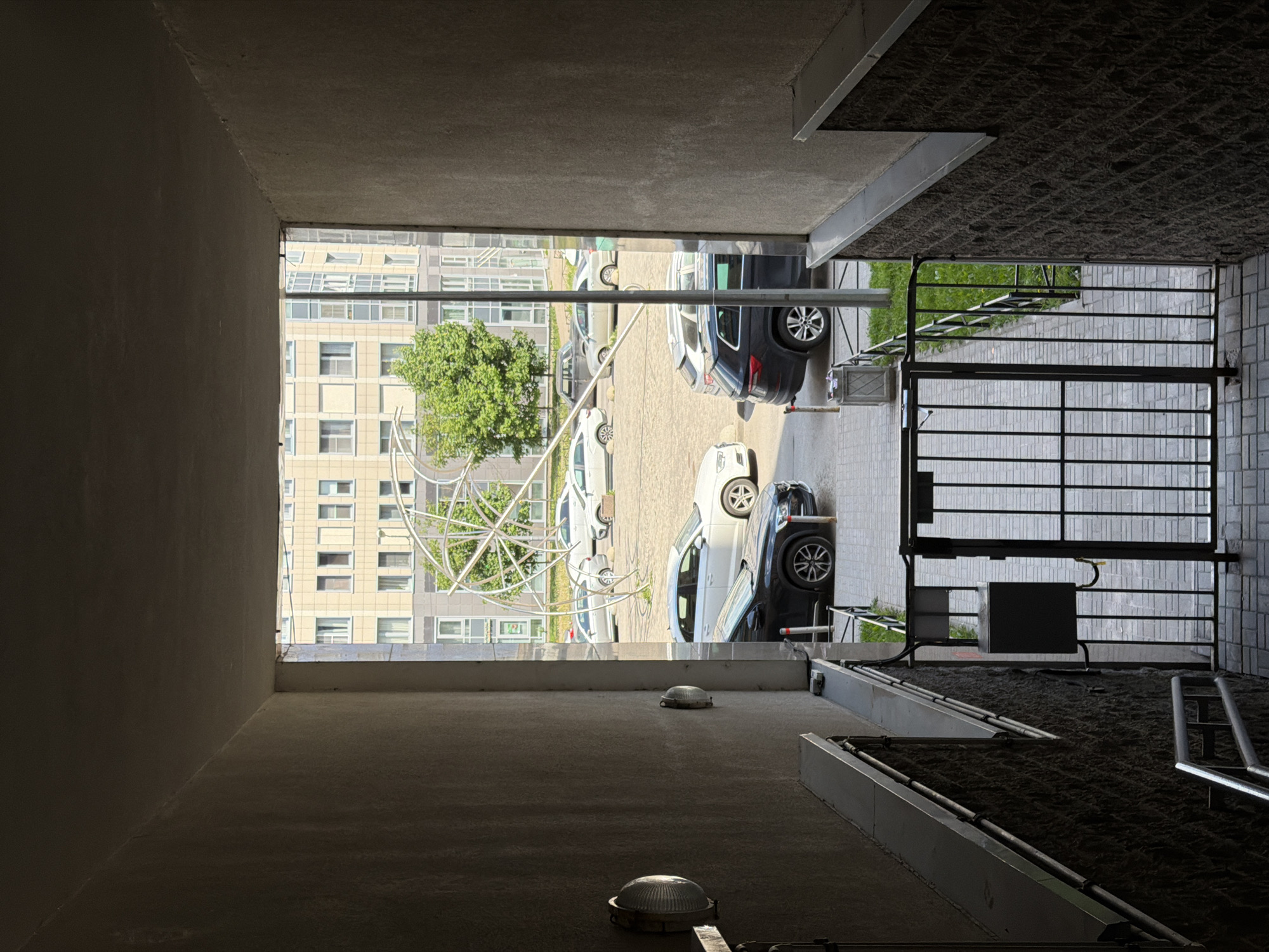 The image depicts an urban outdoor scene framed by a dark, concrete underpass or covered structure. The perspective is from within the shadowed interior, looking out into a brightly lit exterior.
In the foreground, the concrete floor, walls, and ceiling of the underpass are visible. Two round, ribbed light fixtures are mounted on the ceiling. To the right, a textured, dark ground surface, possibly a ramp or path, with metal rails runs along the side.
Transitioning to the exterior, a black metal gate with horizontal bars stands on the right, likely controlling access to the area. Nearby is a black control box and associated wiring, suggesting an automatic barrier system. A low, green fence runs along a sidewalk or path.
The mid-ground reveals a paved parking lot with several parked cars, predominantly light-colored sedans and SUVs, with one dark SUV visible on the right.
In the background, a large, multi-story apartment building with numerous windows and a light-colored facade occupies the left side of the frame. To its right, a vibrant green tree is noticeable, partially encircled or accompanied by a distinctive, large metallic sculpture resembling a spherical or globe-like structure made of interconnected rings.
The strong contrast between the dark interior and the sunlit exterior indicates a clear, sunny daytime. No people or visible text are present. The architecture and urban environment are consistent with a city setting like Saint Petersburg, Russia.