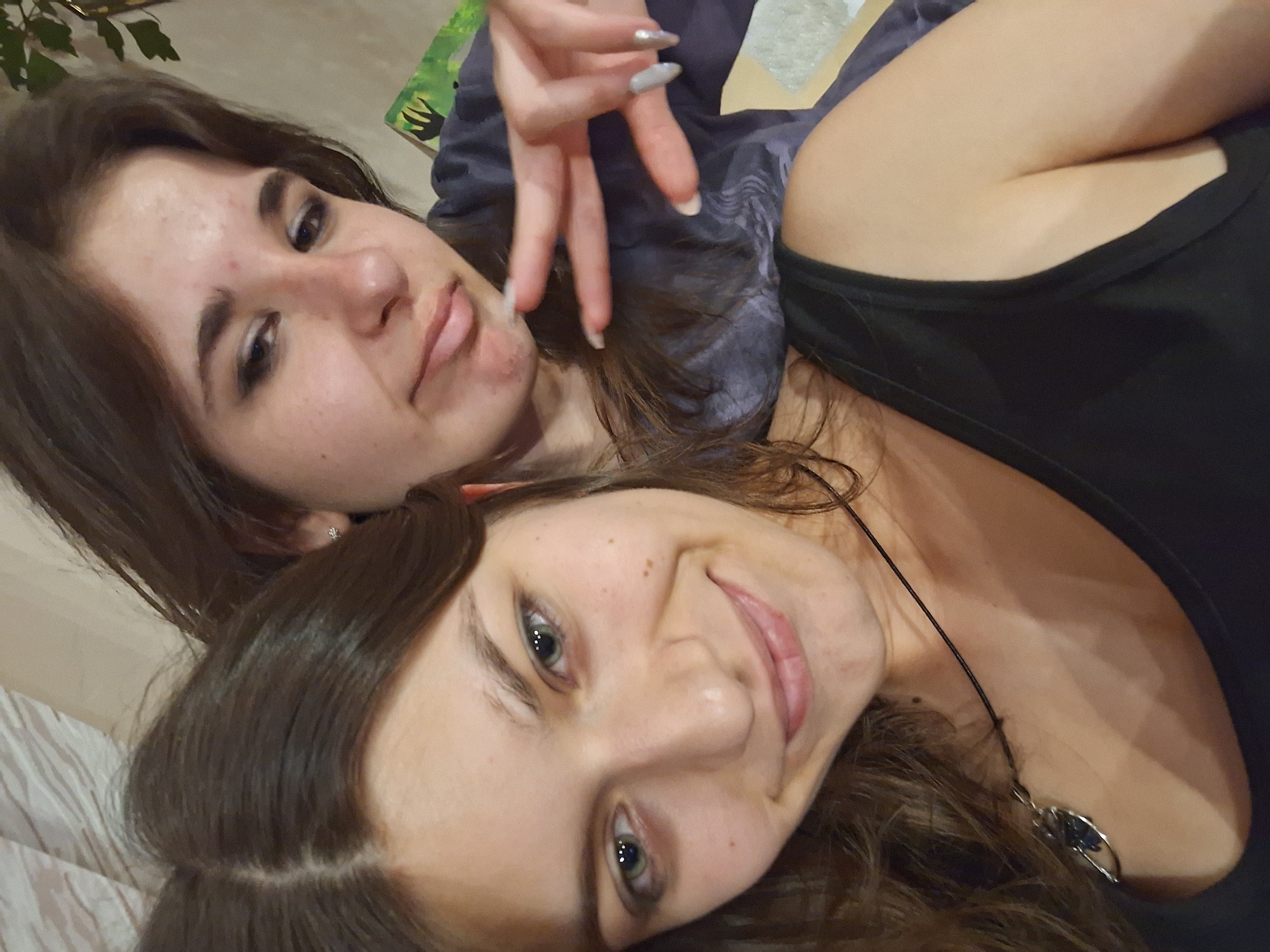 Two young women are lying close together, taking a selfie. The woman on the top has shoulder-length brown hair, fair skin with some acne, and is making a silly face. Her fingernails are long and painted silver. She is wearing a gray shirt. The woman on the bottom has shoulder-length brown hair, fair skin with freckles, green eyes, and is smiling. She is wearing a black tank top and a necklace with a silver pendant. The location is not identifiable from the image.