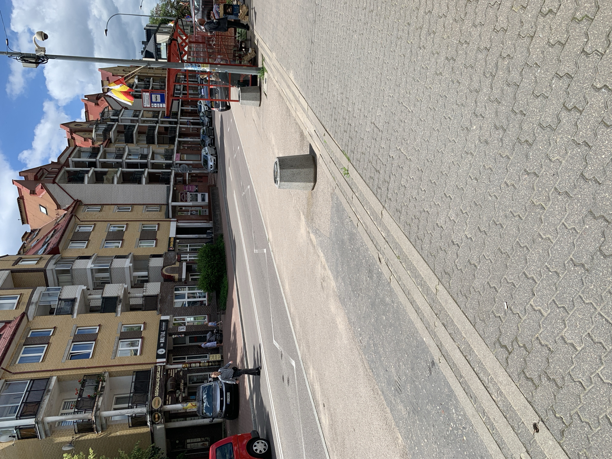 A street scene in Białystok, Poland, features a row of multi-story buildings with yellow facades and red tile roofs. Several businesses are visible on the ground floor, including "REGIONALNE SPECIALY" and "THE BRUTAL BARBER SHOP." A man in a checkered shirt walks across the street, and other pedestrians are seen on the sidewalk. Cars are parked along the street and a bus stop shelter with flags is present. The street is paved with asphalt, and the sidewalk is made of interlocking concrete pavers. A concrete trash receptacle stands on the sidewalk. The sky is partly cloudy.