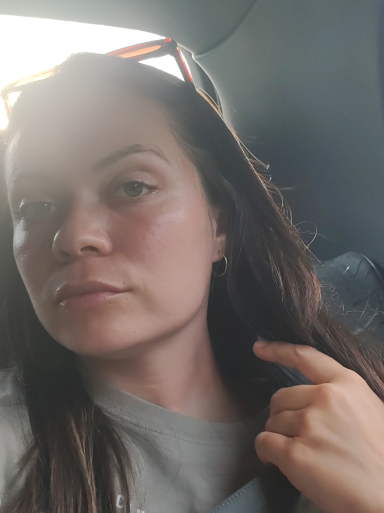 A woman is captured in a selfie taken inside a vehicle, presumably in Garevaya, Russia. She looks directly at the camera with a neutral expression. Her long, dark brown hair frames her face, with strands catching the light. She has green eyes and is wearing simple silver hoop earrings. A pair of brown-framed sunglasses rests on top of her head.
A strong, bright light source from the upper left of the frame creates a significant lens flare, obscuring part of her forehead and hair. This bright illumination highlights her face and gives her skin a natural, possibly slightly dewy, appearance. She is wearing a light gray t-shirt. Her right hand is positioned near her shoulder, with fingers lightly touching her hair or collar. The background reveals the dark gray interior of the vehicle. The intense glare suggests it is daytime and sunny. No text is visible.
