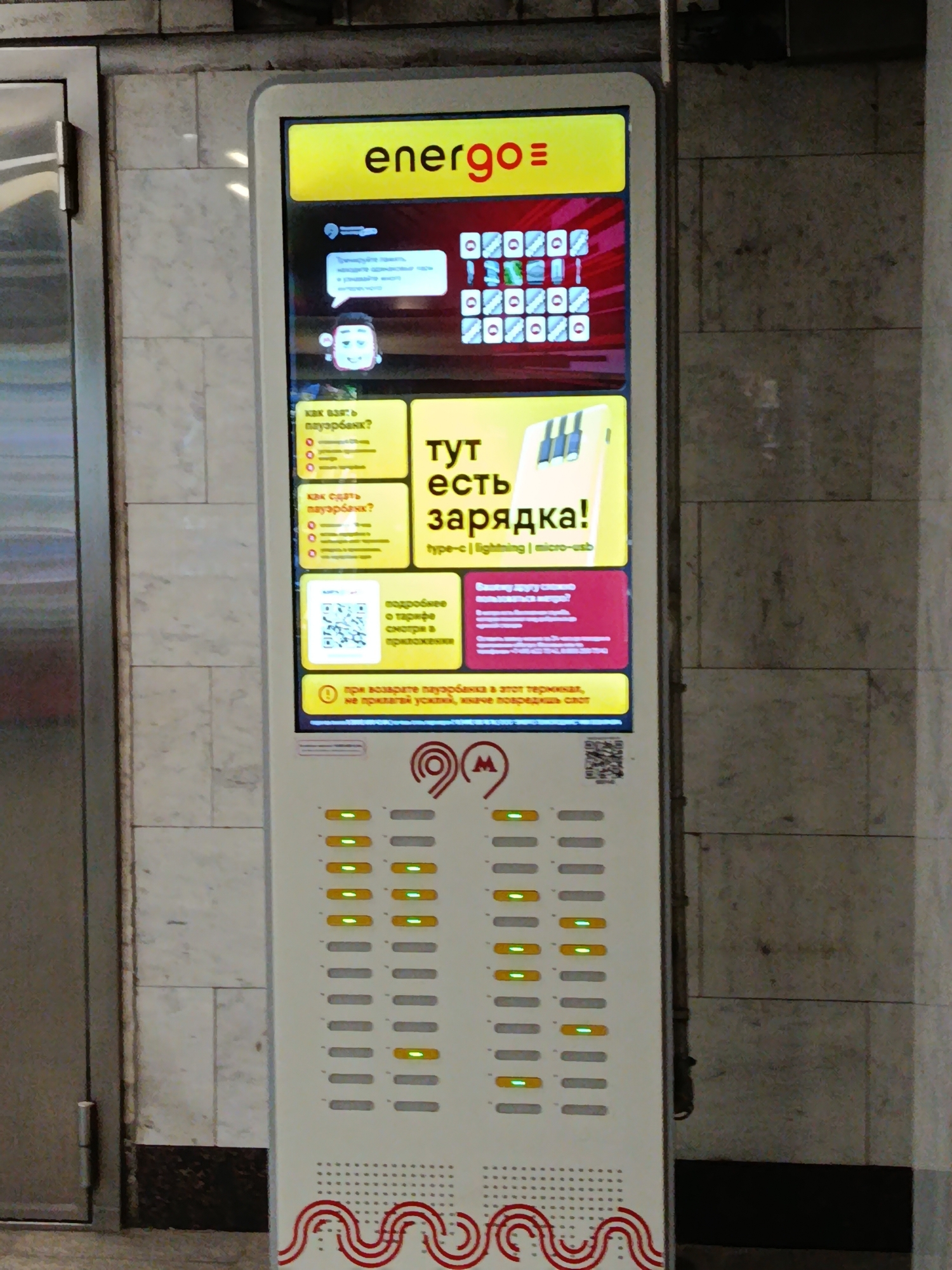 A tall, vertical advertising and charging station is shown in Moscow, Russia, in front of a tiled wall. The station has a bright yellow top section with the word "energo" and a logo. Below this, there is a section with images of phone chargers and text in Russian. The central part of the station displays various charging slots with green indicator lights, suggesting some are in use. The bottom features the Moscow Metro logo. The station appears to be a public charging kiosk.